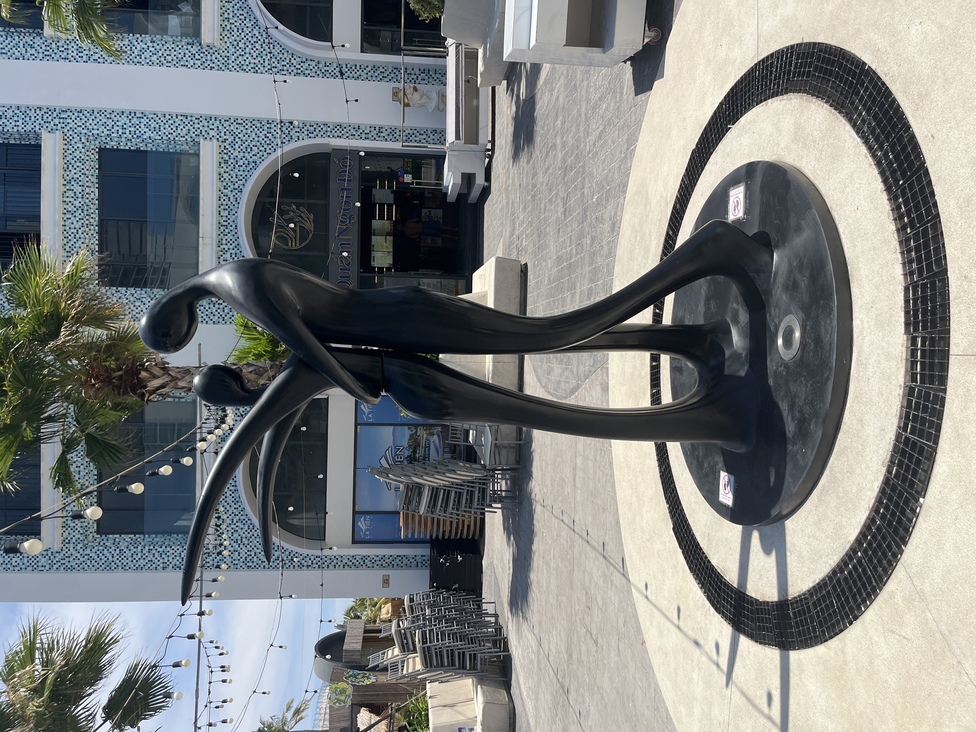 The image shows a black, abstract sculpture of two elongated figures in an embrace, likely made of metal. The sculpture is situated in front of a building with distinctive blue and white mosaic-tiled facade, the building signage reads "QUAN NGON DO" and "LA TIEN". The ground features a circular pattern of light concrete bordered by black mosaic tiles. Stacks of metal chairs are visible in the background. Palm trees are located on the left side of the image. The location is likely in Bac Nha Trang Ward, Vietnam based on the provided building sign. The scene appears to be a public space or outdoor area associated with the restaurant.