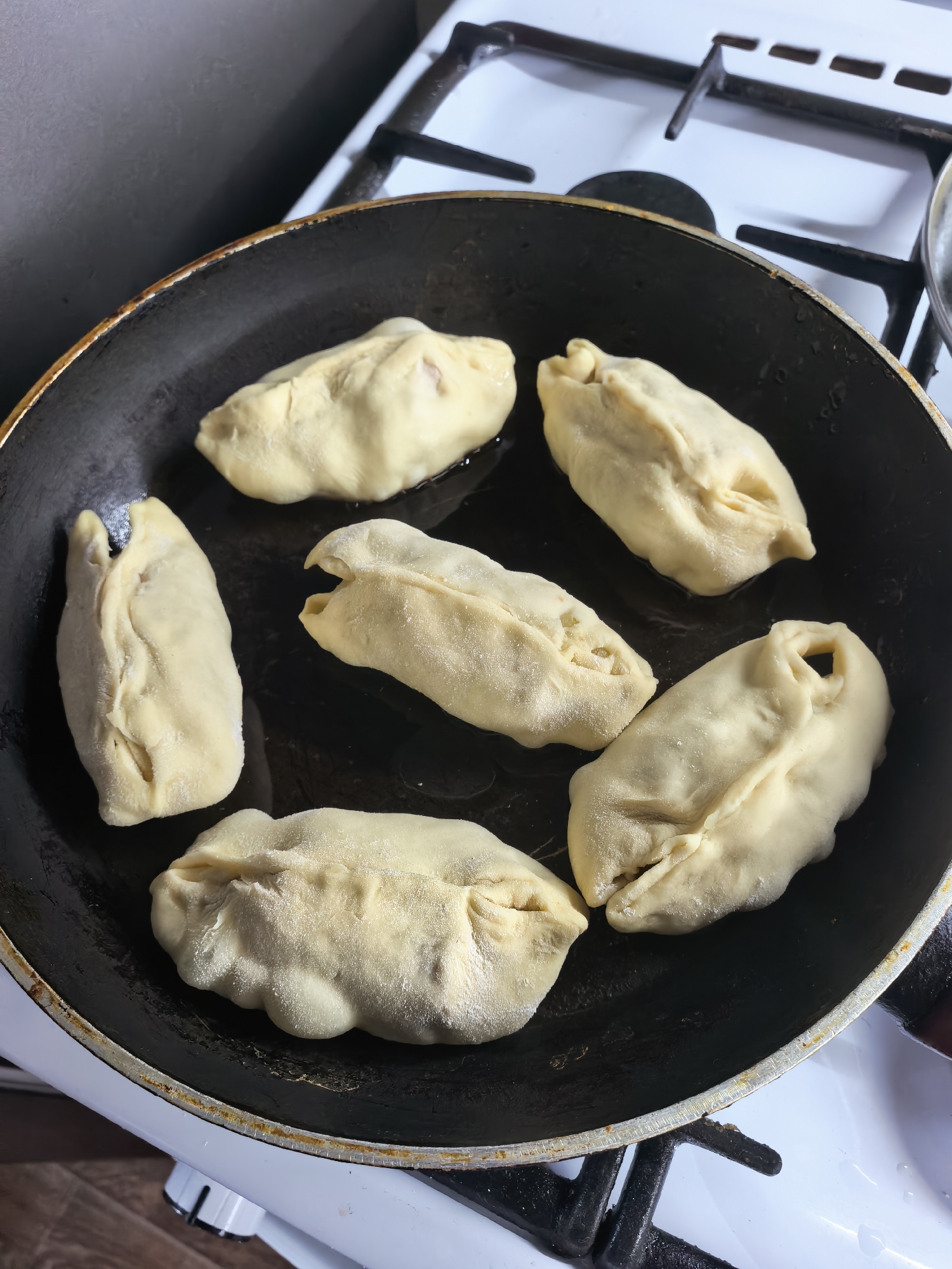 Six uncooked dumplings or pastries are arranged in a black frying pan, resting on a white gas stovetop with black grates. The dumplings are light beige, appear to be made of dough, and show visible folds and pleats, suggesting they are handmade. Some residual flour is visible on their surfaces, and a small amount of oil or liquid is present at the bottom of the pan around them. The pan itself appears well-used, with some discoloration or wear along its rim. The stovetop is clean, featuring a standard design. The scene depicts the initial stage of cooking these items in a domestic kitchen setting, likely in Sterlitamak, Russia. No people are visible, and there is no discernible text. The background consists of a dark, plain wall or surface.