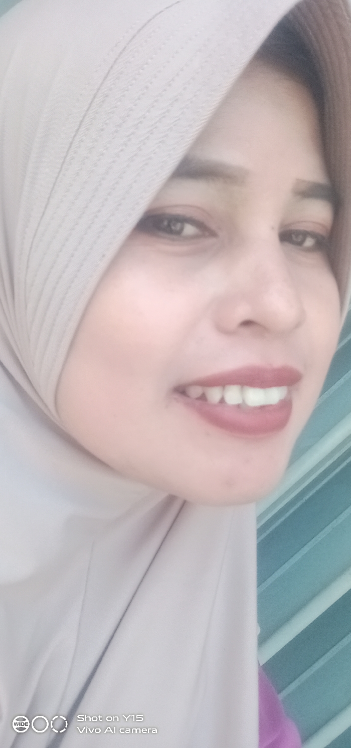 A vertical close-up photograph, purportedly taken in Gending, Indonesia, depicts an adult female. The subject's face is central, partially framed by a light beige head covering (hijab) that features textured, ribbed fabric on its upper section. Her eyes are dark, eyebrows are defined, and lips are colored with reddish-brown lipstick. Her mouth is open in a smile, revealing both upper and lower teeth. The background is out of focus, showing a light blue or teal corrugated or slatted surface to the right of the subject's head, and a small portion of purple fabric in the lower right corner. A "Shot on Y15 Vivo AI camera" watermark is visible in the bottom left corner.