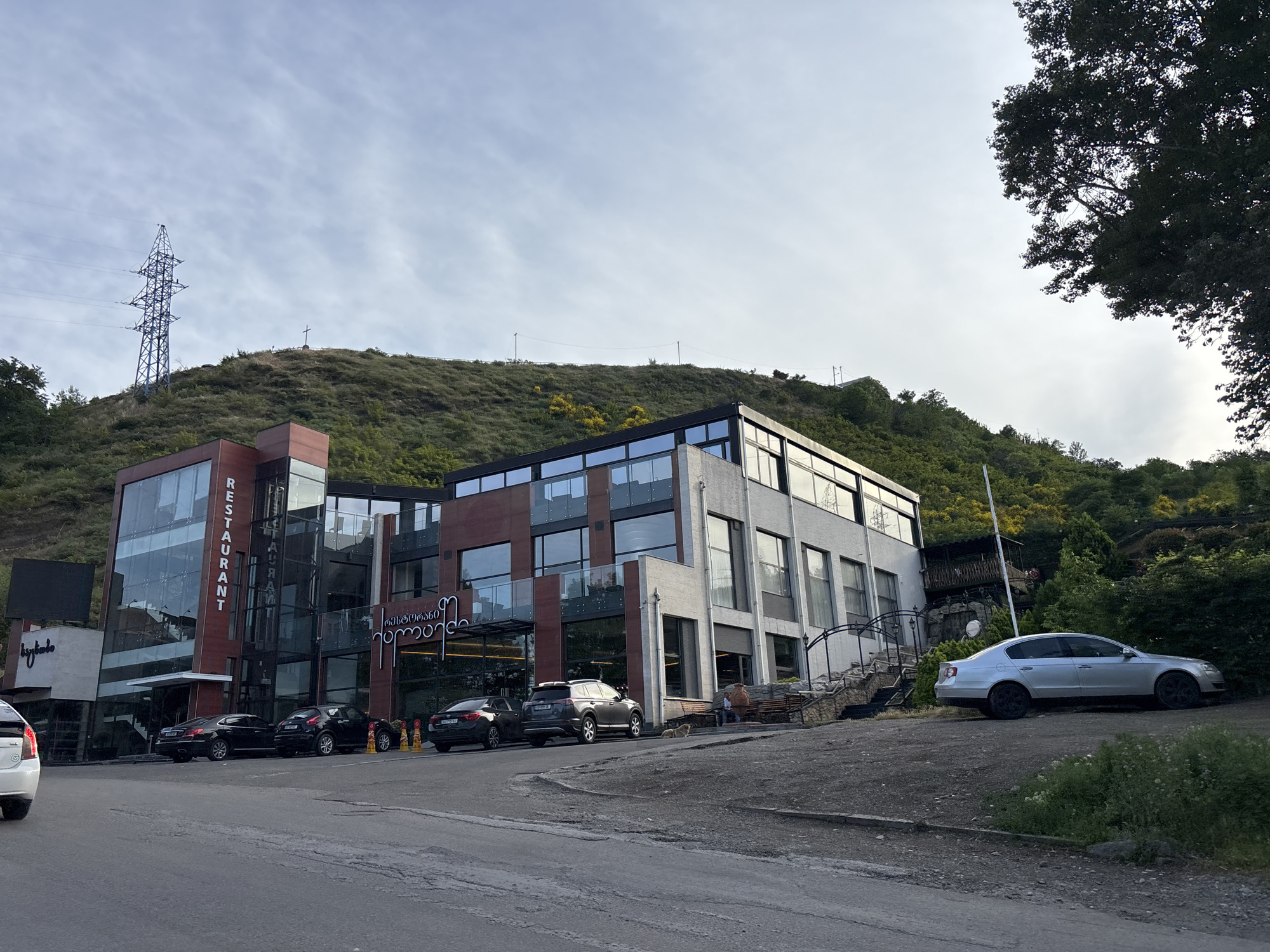 The image shows a modern multi-story building with large glass windows, situated on a hillside in Tbilisi, Georgia. The building appears to be a restaurant or commercial establishment, with signage visible in both English ("RESTAURANT") and Georgian script. Several cars are parked in front of the building, including a silver sedan, a dark SUV, and two dark sedans. The hillside behind the building is covered with greenery and trees, and a large power transmission tower is visible in the distance on the left, along with a cross on the hilltop. The sky is overcast with clouds. In the foreground, a paved road with some wear and tear leads past the building. A small dog is lying down on the ground near a bench outside the building on the right side.