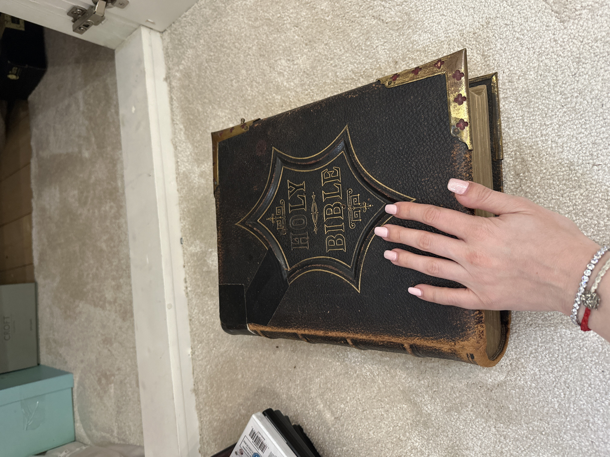 A large, antique-style Holy Bible rests on a light-colored, flecked carpeted floor. The book features a dark, textured cover with ornate gold-embossed text reading "HOLY BIBLE" within a decorative frame, and prominent metal corner embellishments secured with small, floral-shaped studs. The spine and edges of the book exhibit wear and discoloration, showing a brownish patina. Gilded page edges are visible. A human left hand, with light pink nail polish, is positioned on the book's cover. The wrist displays multiple bracelets: a red string bracelet, a silver-toned tennis bracelet, and another thin bracelet with a metallic charm. To the left of the Bible, a partial stack of white-cased optical discs is visible on the carpet. In the background, a white threshold separates the foreground carpet from another room or area. Beyond the threshold, a light blue box, a box partially labeled "CROFT," and a dark box are discernible. A white door frame with a silver hinge is visible on the far right. The specific location within the city of London, United Kingdom, cannot be determined from the provided visual information.