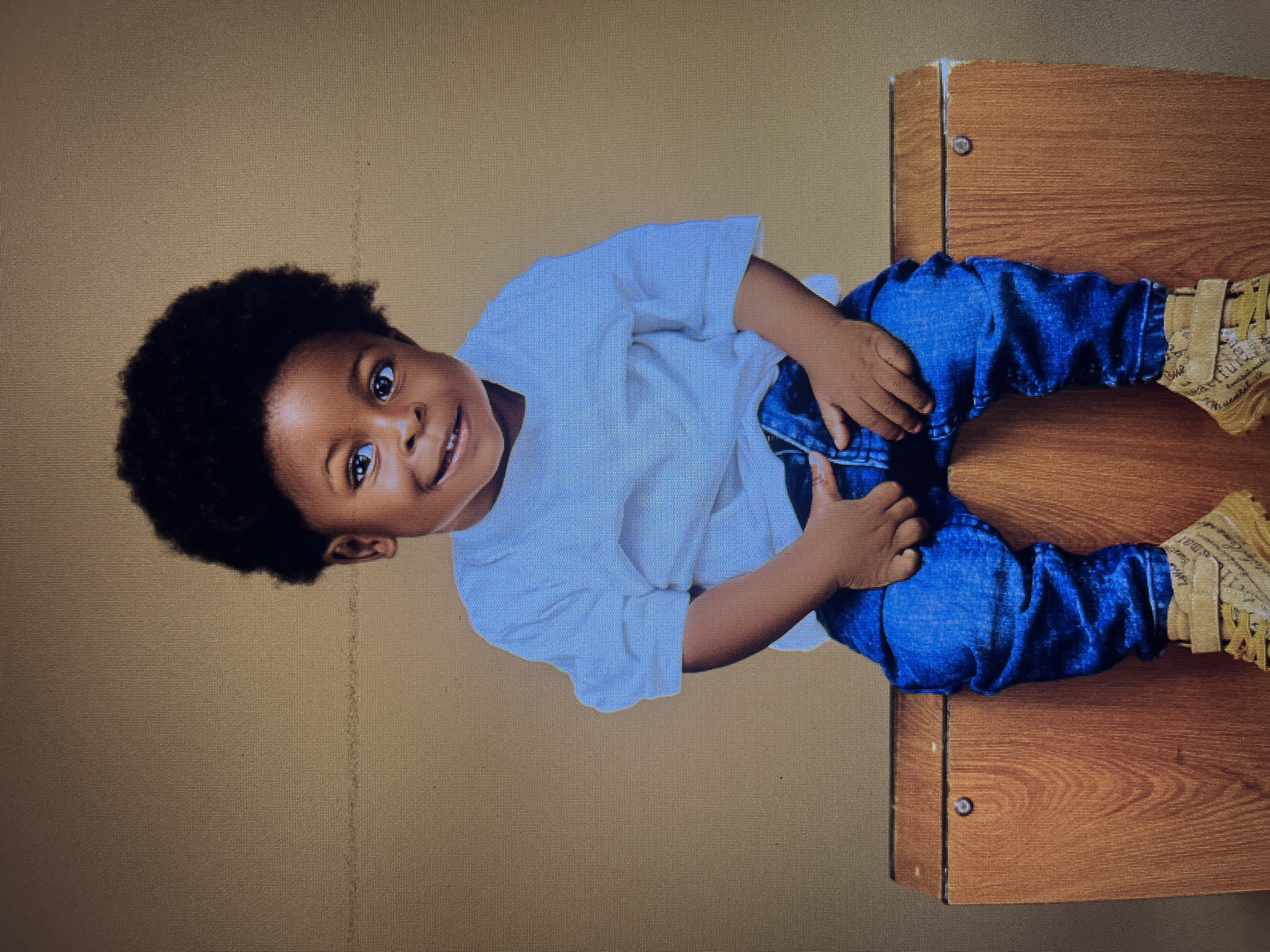 A young Black child, likely a toddler, is seated on a wooden surface against a plain, textured brown backdrop. The child has dark, curly hair and is wearing a white t-shirt and blue jeans. Their hands are resting on their lap, and they are looking directly at the camera with a slight smile. The wooden surface appears to be a piece of furniture, possibly a bench or cabinet, with visible grain and some metal hardware. The overall setting suggests a professional studio portrait. There is no indication of the location being in Gura Top, Nigeria, or any other specific city.