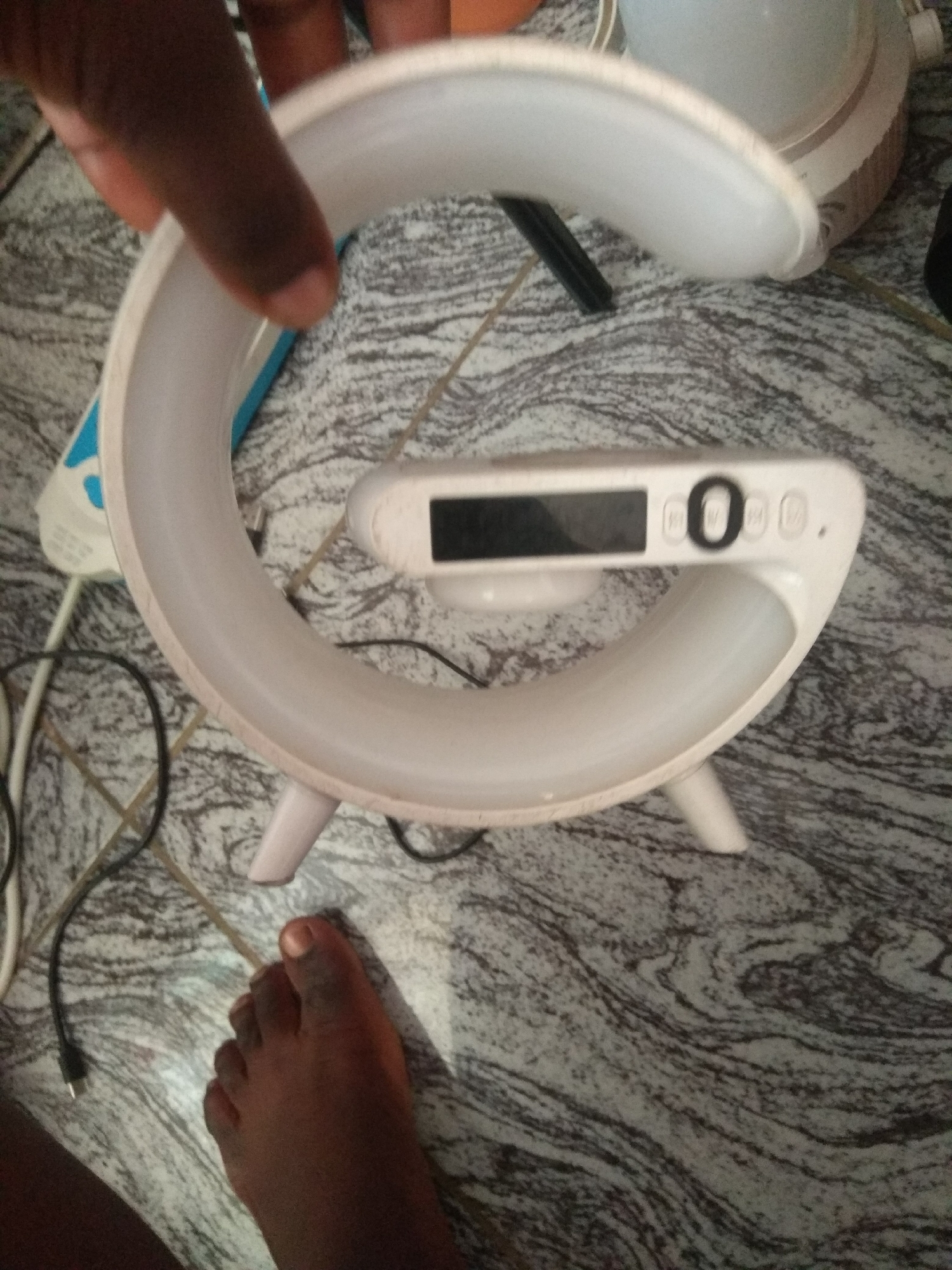 This media file features a close-up shot of a white, crescent-shaped object, possibly a lamp or a unique alarm clock, resting on a patterned floor. The object has a digital display showing what appears to be a "0" and other unreadable symbols or buttons. The device has a modern, minimalist design with subtle LED lighting visible. A black wire is connected to the device, extending out of frame, and another white wire is also visible.
In the foreground, a person's dark-skinned foot is visible, with toes slightly curled. A dark-skinned hand is also present in the upper left corner, partially obscuring a blue and white object in the background, which could be another electronic device. The floor appears to be tiled with a marbled pattern of white and dark grey or black lines. The lighting suggests an indoor setting, and the overall impression is casual and everyday. The location is identified as Jalingo, Nigeria. There are no discernible cues about the time of day or weather conditions. No explicit emotions are conveyed, beyond a sense of everyday life.
