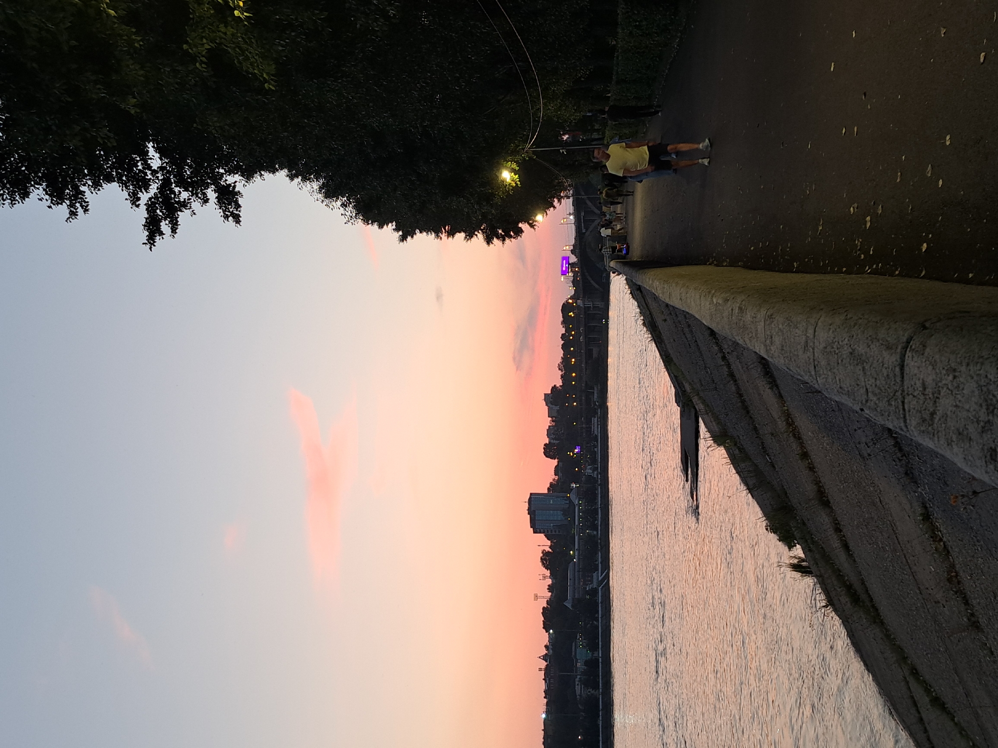 This is a vertical, medium-shot, outdoor photograph taken at dusk along the Angara River embankment in Irkutsk, Russia. The sky is a gradient of light blue at the top, fading to soft pinks and oranges towards the horizon, with a few wispy clouds visible.
On the left side of the image, the wide, calm Angara River occupies the lower left quadrant, its surface reflecting the colors of the sky. A concrete embankment with some sparse vegetation lines the riverbank.
To the right of the river is a paved walkway. Several people are walking or jogging along the path. In the foreground, a man wearing a yellow t-shirt and dark shorts is jogging. Further down the path, other individuals are visible, some walking in groups. The walkway is lined with trees on the right side, casting shadows and obscuring the view beyond. Streetlights along the walkway are illuminated, providing a warm glow.
In the distance, across the river, is a city skyline with buildings of various sizes, including a taller, modern-looking building. A bridge is visible spanning the river in the mid-ground, and lights from the city can be seen twinkling. The overall atmosphere is serene and tranquil, indicative of an evening stroll.