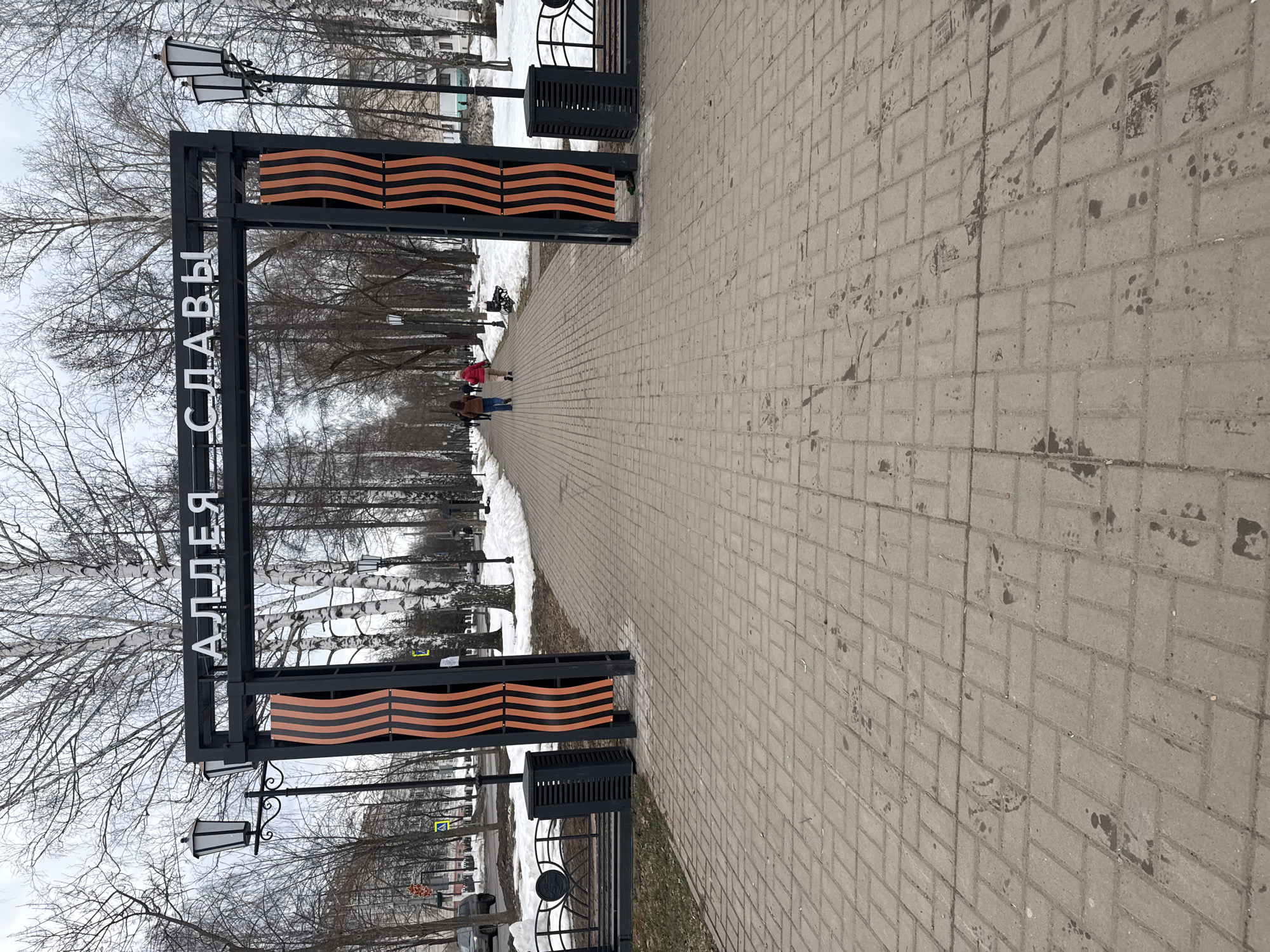 Here is a description of the image:
The image shows a brick-paved pathway in a park or public space. An arched gateway, decorated with black and orange horizontal stripes and Russian lettering (reading "Alley of Glory"), spans the entrance to the path. Two people are walking along the path toward the middle distance. There is snow on the ground to the sides of the path. Several leafless trees are visible. There are lampposts on either side of the pathway. The image location is in the city of Chaikovsky, Perm Krai, Russia.