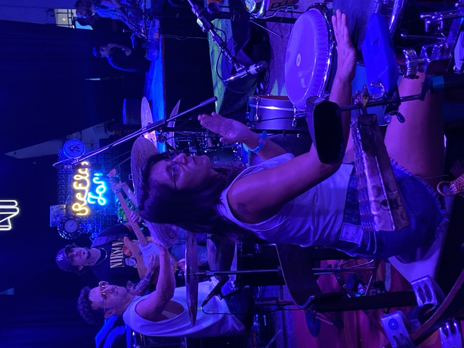 The image shows a live music performance occurring inside a venue, located in Buenos Aires, Argentina. The environment is dimly lit with predominant blue and purple stage lighting. On the back wall, a yellow-orange neon sign displaying stylized text, possibly "ReElect.Ton" or "ReElect.ion," is visible alongside a dollar sign symbol.
In the foreground, a woman with dark, shoulder-length hair and glasses is seated on a stool, performing percussion. She wears a white tank top, light-colored denim shorts, and a blue wristband on her right wrist. Her right hand rests on a large percussion drum, likely a conga, while her left hand is positioned near a mounted cowbell and a blue jam block.
To the left and slightly behind her, a man with short, curly hair and glasses is seated, facing a drum kit. He wears a white sleeveless top and holds drumsticks, with a cymbal visible in the mid-ground.
Further back and centrally, a third man stands, playing an electric guitar with a light-colored body. He wears a dark baseball cap with a white "NY" logo and a dark t-shirt featuring a "NIRVANA" graphic.
Multiple microphone stands, various cables, and additional musical equipment are present across the stage area. In the background, other individuals are dimly visible, suggesting additional band members or an audience beyond the immediate performance area.