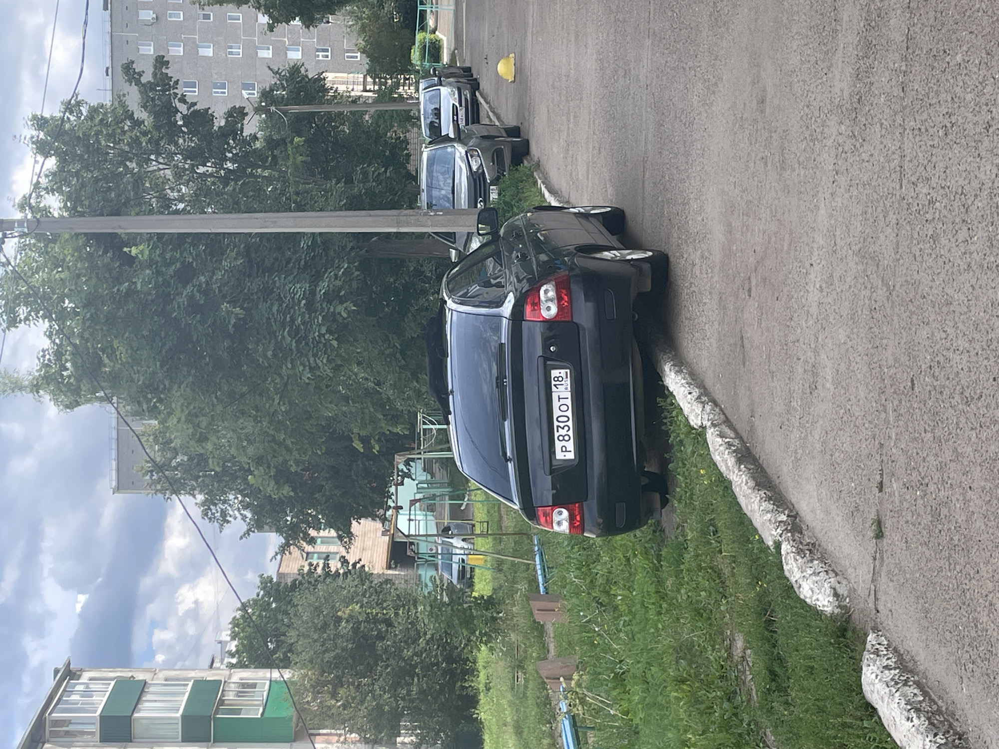 The image depicts a street scene in Sarapul, Russia, under a cloudy sky. In the foreground, a black sedan is parked on the side of a paved road, its rear end facing the camera. The car has a Russian license plate that reads "P 830 OT 18 RUS". To the left of the car is a patch of overgrown green grass and a concrete curb. A children's playground with swings and metal structures is visible in the background, along with more greenery and trees. Beyond the playground, residential buildings are present. Further down the street, several other cars are parked in a line. A yellow traffic cone is also visible on the right side of the road. Utility poles with wires are scattered throughout the scene. The environment appears to be a typical urban residential area.