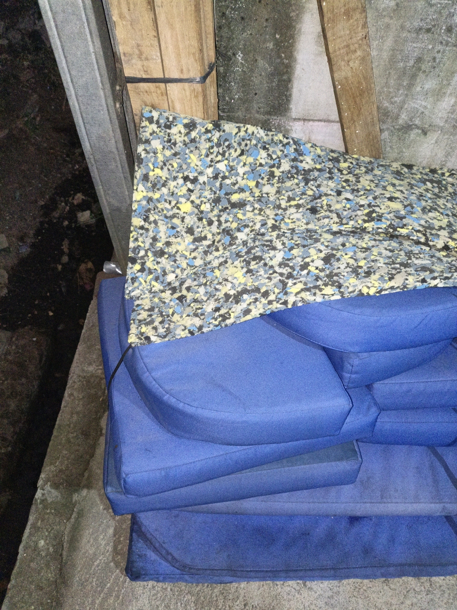 The image shows a pile of blue cushions stacked on top of each other, with a piece of multi-colored foam padding on top. The cushions appear to be made of a durable fabric and are slightly worn. The foam padding is a mix of blue, yellow, and black pieces. The scene is outdoors, with a rough concrete surface and dirt visible in the background. There are also wooden planks and a metal beam present, suggesting a construction or storage area. The lighting is dim, and there are no people visible in the image. The location in Cikal, Indonesia, is not directly identifiable from the image itself, but the overall scene suggests an outdoor storage or disposal area within a semi-urban or rural setting.
