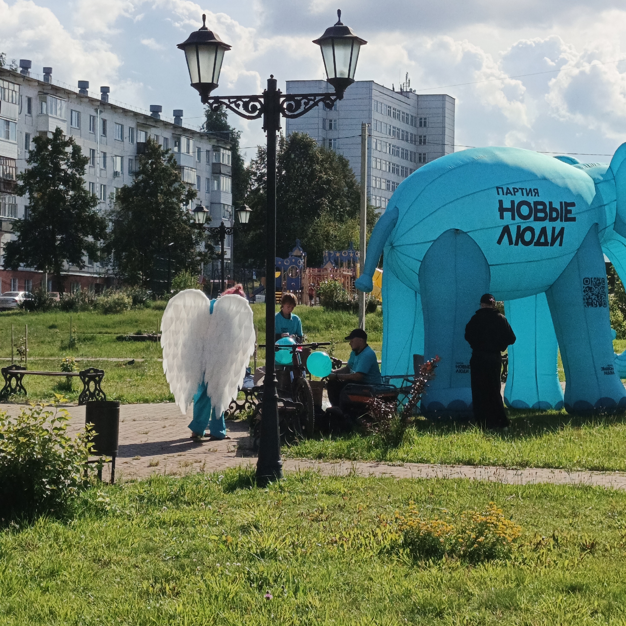 The image depicts an outdoor scene in the city of Chaikovsky, Russia, likely in a park or public square. Dominating the right side of the frame is a large, inflatable blue elephant, part of an advertisement for "ПАРТИЯ НОВЫЕ ЛЮДИ" (Party New People). The text is clearly visible in black lettering on the elephant's body. To the left of the elephant, a person in a black robe stands facing the inflatable.
In the foreground left, a person dressed in a blue costume with large white angel wings is standing on a paved walkway. They are facing away from the camera. Further to the left, a dark metal trash receptacle is situated near a park bench.
In the middle ground, two individuals in matching blue t-shirts are interacting with a bicycle and a bright blue balloon. One person is seated on a bench, while the other stands. A second, smaller lamppost with two lamps is visible behind them, and a larger lamppost with a single lamp is in the mid-left of the frame.
In the background, typical apartment buildings of a Russian city are visible, with trees and a children's playground adding to the urban park setting. The sky is partly cloudy. The overall impression is of a public event or promotional activity taking place outdoors.
