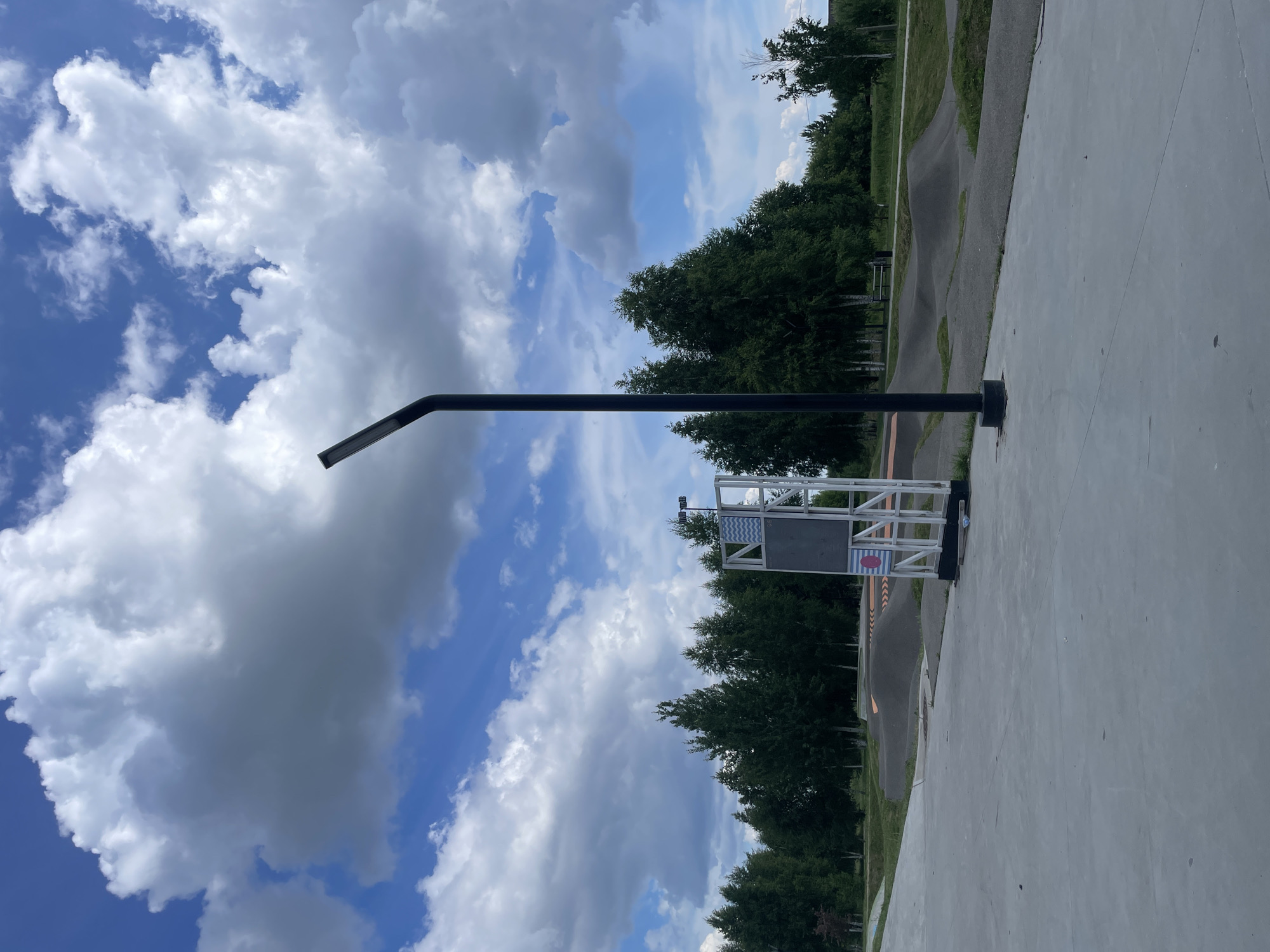 An outdoor shot shows a pump track for bicycles and skateboards in Laishevo, Russia. The pump track is composed of a series of dirt mounds and banked turns, with a paved area in the foreground. A tall, black lamppost stands to the right of the frame, with a white metal structure behind it displaying a blue and white chevron pattern and a pink circle. In the background, a dense line of green trees creates a natural border, and a bright blue sky with fluffy white clouds stretches above. The overall scene is one of a recreational park in a natural setting.
