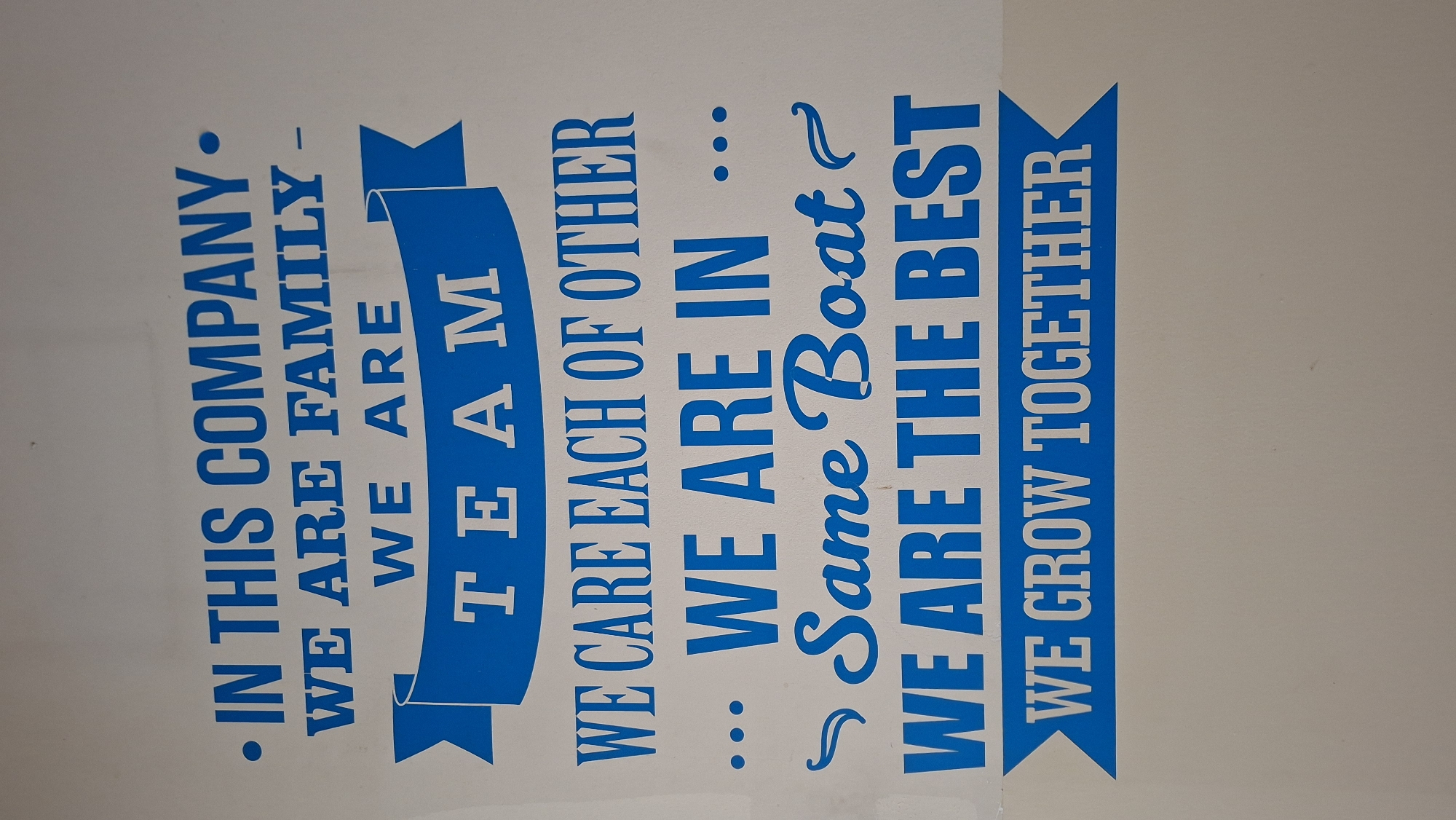 The image displays a sign with blue text on a white background. The sign contains several lines of text. The top line reads ". IN THIS COMPANY ". The second line reads "WE ARE FAMILY -". The third line reads "WE ARE". The fourth line, within a blue banner, reads "TEAM". The fifth line reads "WE CARE EACH OF OTHER". The sixth line reads "... WE ARE IN ...". The seventh line, in a decorative script font, reads "Same Boat". The eighth line reads "WE ARE THE BEST". The ninth line, within a blue banner, reads "WE GROW TOGETHER". The sign is mounted on a plain white wall. The image does not provide any information about its location in Palembang, Indonesia.