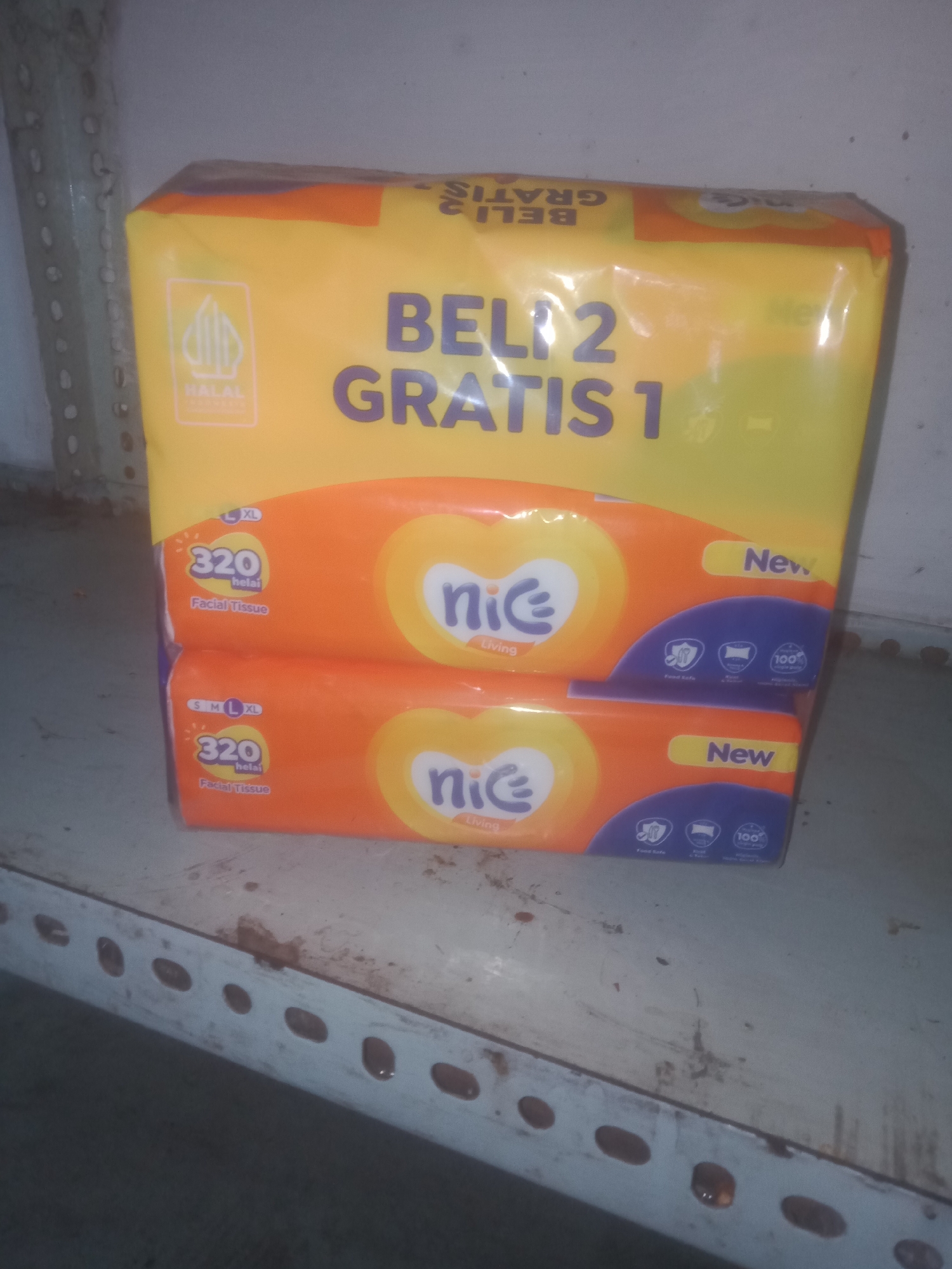 Two packs of Nice brand facial tissues are stacked vertically on a metal shelf. The top pack features yellow and orange packaging with "BELI 2 GRATIS 1" (Buy 2 Get 1 Free) printed in large purple letters. A "HALAL" logo is visible on the upper left corner. The bottom pack has similar orange and yellow packaging with a "New" label on the right side. Both packs indicate "320 helai" (320 sheets) and "Facial Tissue" in smaller print. The shelf is weathered and appears to be in an indoor setting, possibly a storage area. The image was taken in Badas, Indonesia.