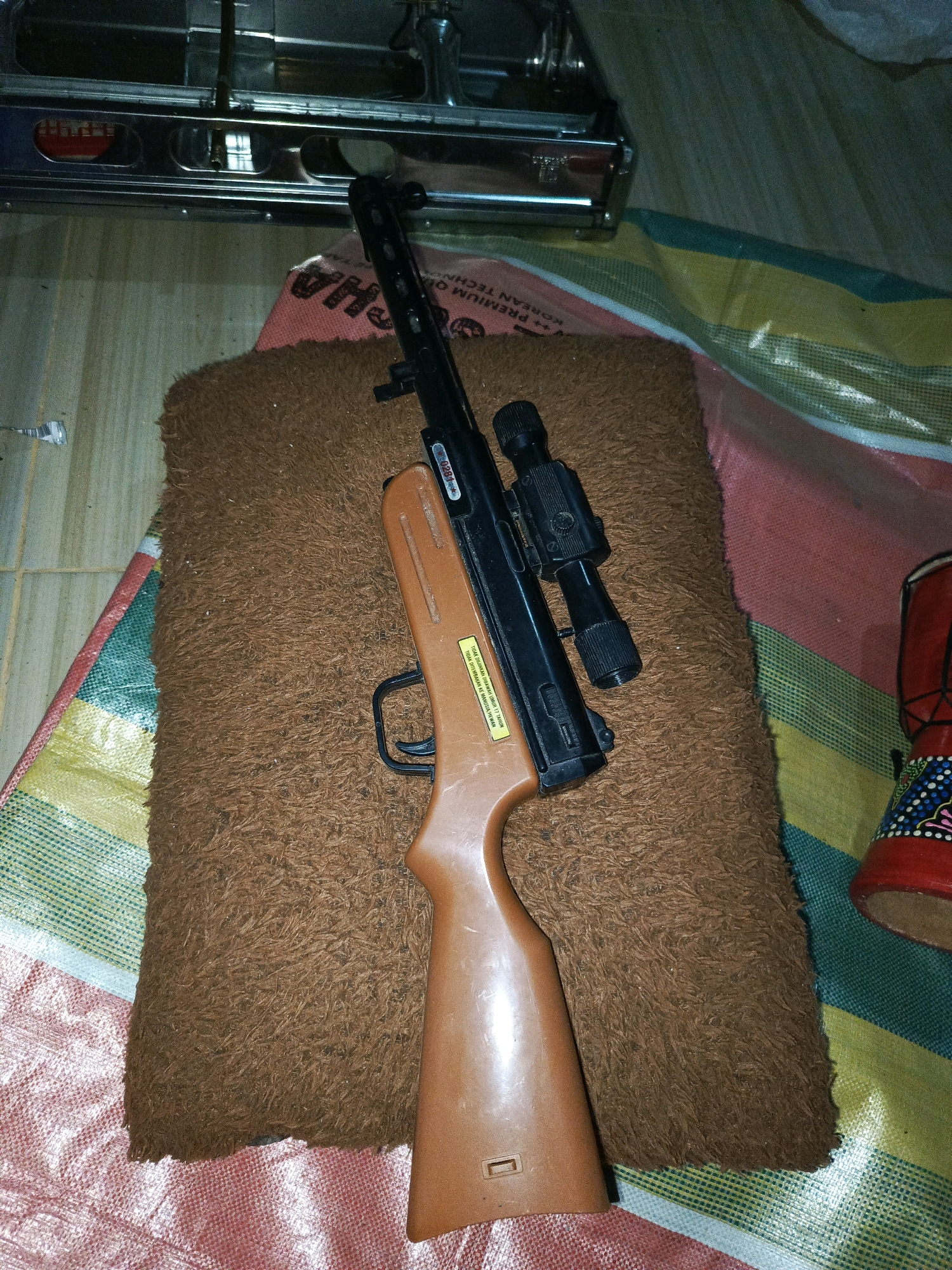 The image shows a rifle with a scope, resting on a brown, furry mat. The rifle is a toy gun, with an orange stock and a black barrel. The scope is also black. The mat is placed on a colorful patterned surface, which appears to be a large plastic sheet or tablecloth. In the background, a metallic object, possibly a piece of furniture or equipment, is visible. The lighting suggests the image was taken indoors in dim conditions. There are no people or animals visible in the image. The setting does not provide any specific clues to its location within Megang Sakti I, Indonesia.