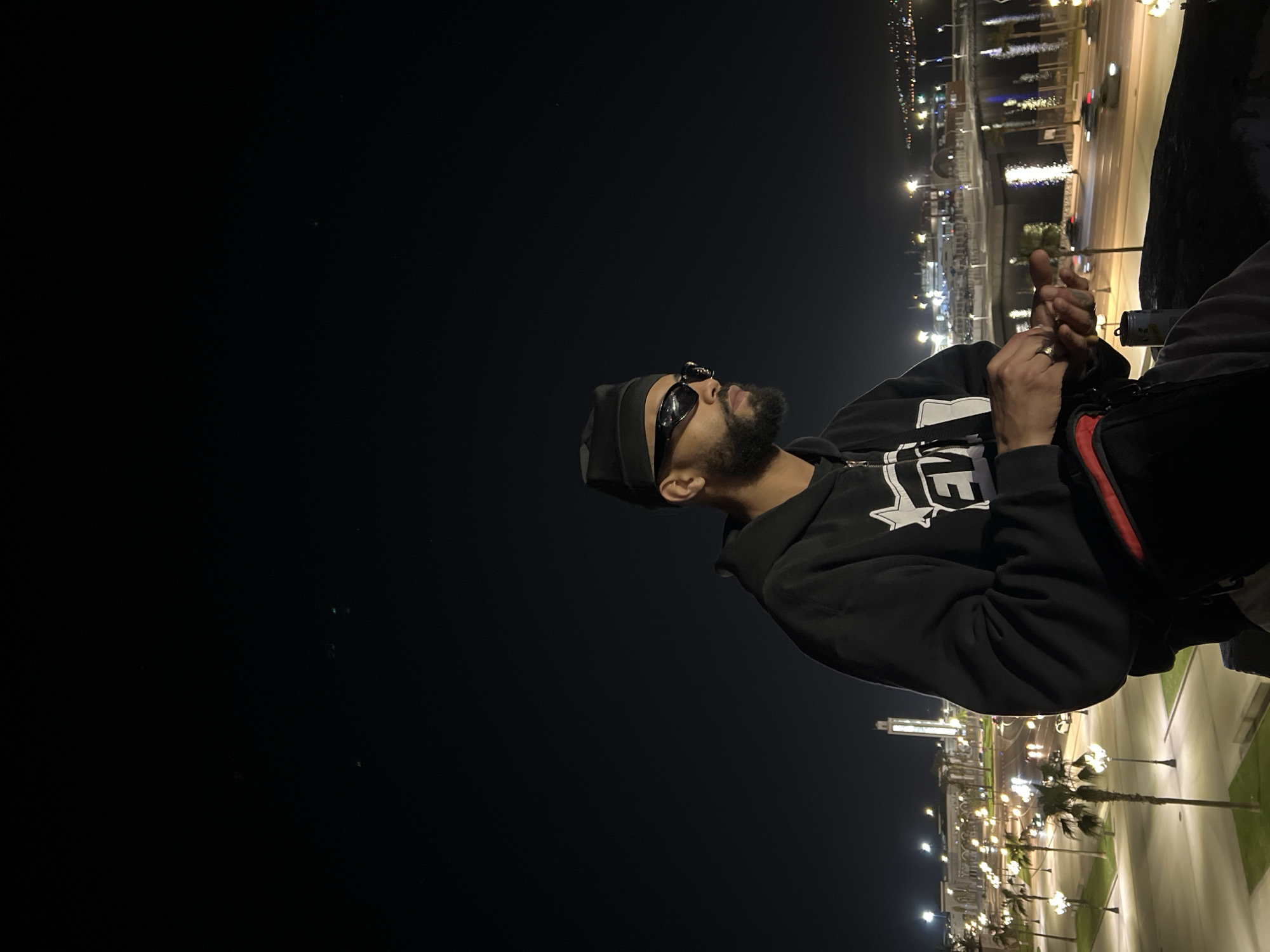The image depicts a nighttime urban scene in Tanger, Morocco, overlooking the city's waterfront. A male individual, characterized by a beard and mustache, is seated in the foreground, facing towards the right side of the frame. He is wearing a black beanie, dark wraparound sunglasses, a black hooded sweatshirt with a partially visible white graphic, and dark trousers. A black fanny pack with a red zipper is strapped around his waist. His hands are positioned centrally, manipulating a small, metallic object. A silver-colored can rests on the surface to his left. A silver-colored ring is visible on his left ring finger.
The background presents an illuminated cityscape. A wide, multi-lane road, lined with numerous palm trees and streetlights, occupies the mid-ground, showing blurred streaks of light from moving vehicles. Beyond the road, a body of water, likely the port or marina, reflects the surrounding urban illumination. Across the water, distant city lights delineate the horizon. A prominent, tall, illuminated square minaret, belonging to a large building, is visible in the left-mid background, consistent with structures near the Tangier port area, such as the Grande Mosquée Mohammed V. The sky is dark and clear.