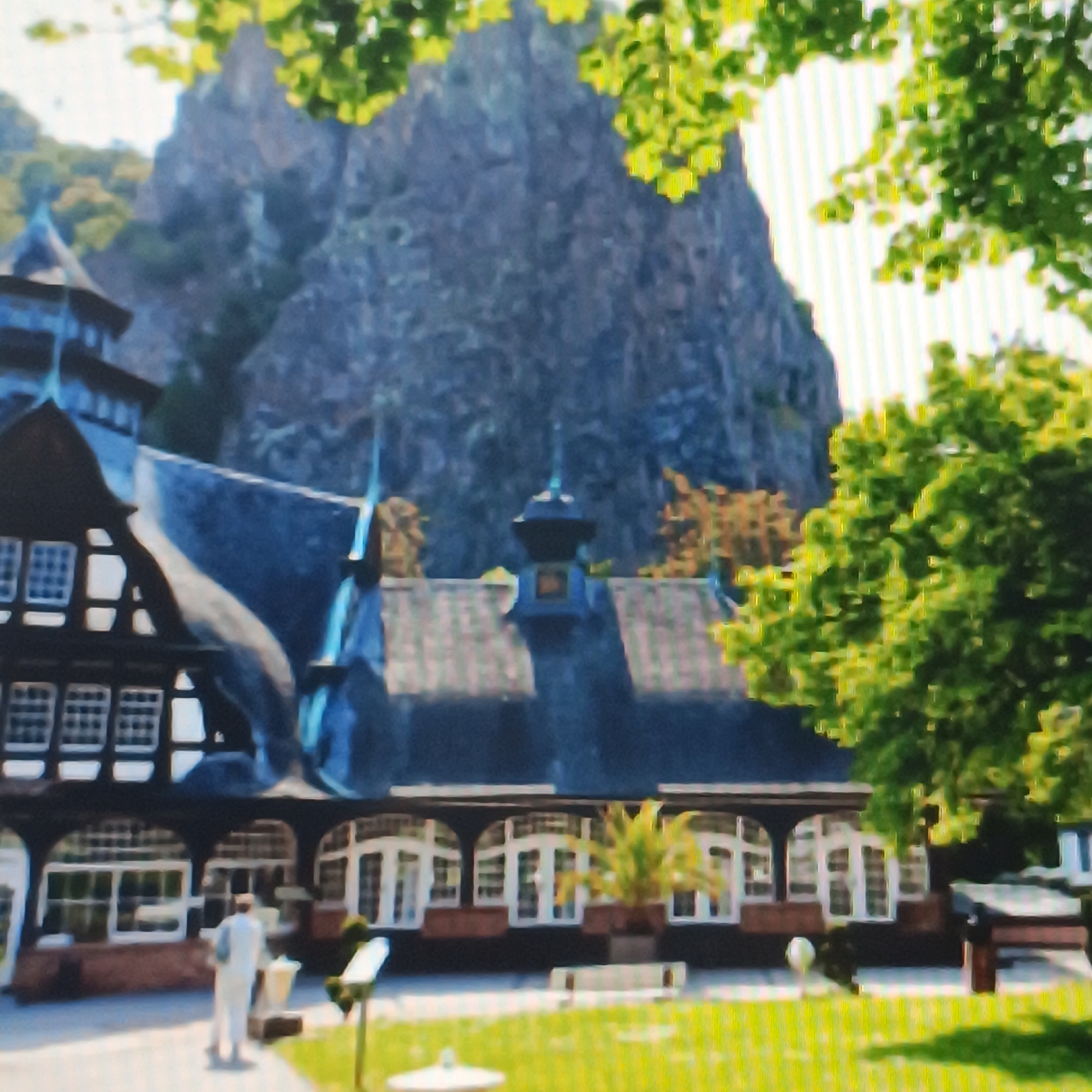 The image shows a building with a dark, angled roof and light-colored windows with dark frames. The building appears to be a hotel or restaurant, with a distinctive architectural style featuring multiple gables and a spire-like structure on one of the roofs. In the background, a large, rocky cliff face rises steeply, partially obscured by lush green foliage. The foreground consists of a manicured lawn with a few scattered objects, including a person in light-colored clothing walking away from the camera. This scene is characteristic of the area around the Opel Villa in Rüsselsheim am Main, Germany, which features historic architecture set against a natural, rugged landscape.