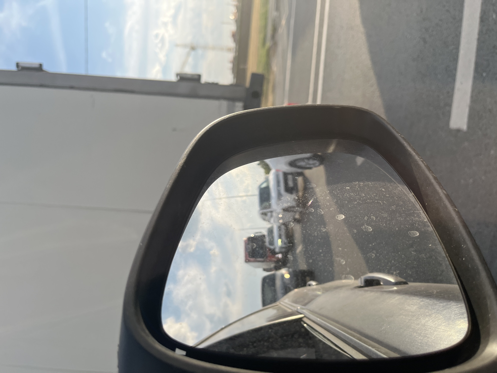 A side mirror reflects traffic congestion on a sunny day, with a cloudy sky overhead. The mirror shows several cars in traffic, including a red truck and a white SUV. In the background, a large white truck and a construction crane are visible. The road appears to be an urban highway with clear lane markings. The image is taken from inside a vehicle, showing the exterior of the car in the foreground on the left side of the frame.