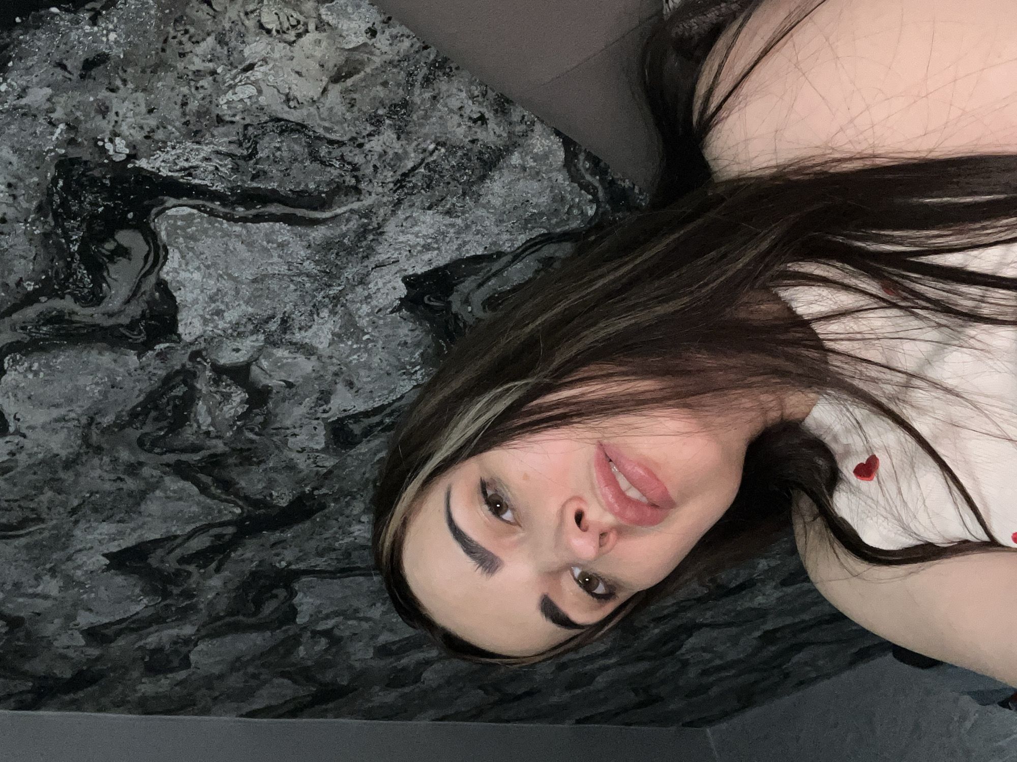 A young woman is the central subject of this close-up, angled photograph. She is depicted lying down or reclining, with her head tilted and facing the camera, giving a direct gaze. Her long, dark brown hair, featuring lighter highlights around her face, flows around her. She has defined eyebrows, visible winged eyeliner, and full lips parted in a subtle, relaxed smile. She is wearing a white top adorned with small red heart patterns, visible on her shoulder and chest.
The background behind her features a distinctive pattern of dark gray, black, and white swirls, resembling marble, granite, or an abstract artistic texture. This patterned surface fills a significant portion of the frame. A solid dark gray area, possibly a piece of furniture or upholstery, is visible in the upper and lower sections of the image, suggesting an indoor setting. The lighting appears soft and even. The woman's expression is calm and composed, indicating a relaxed pose for the photograph. There is no visible text in the image.