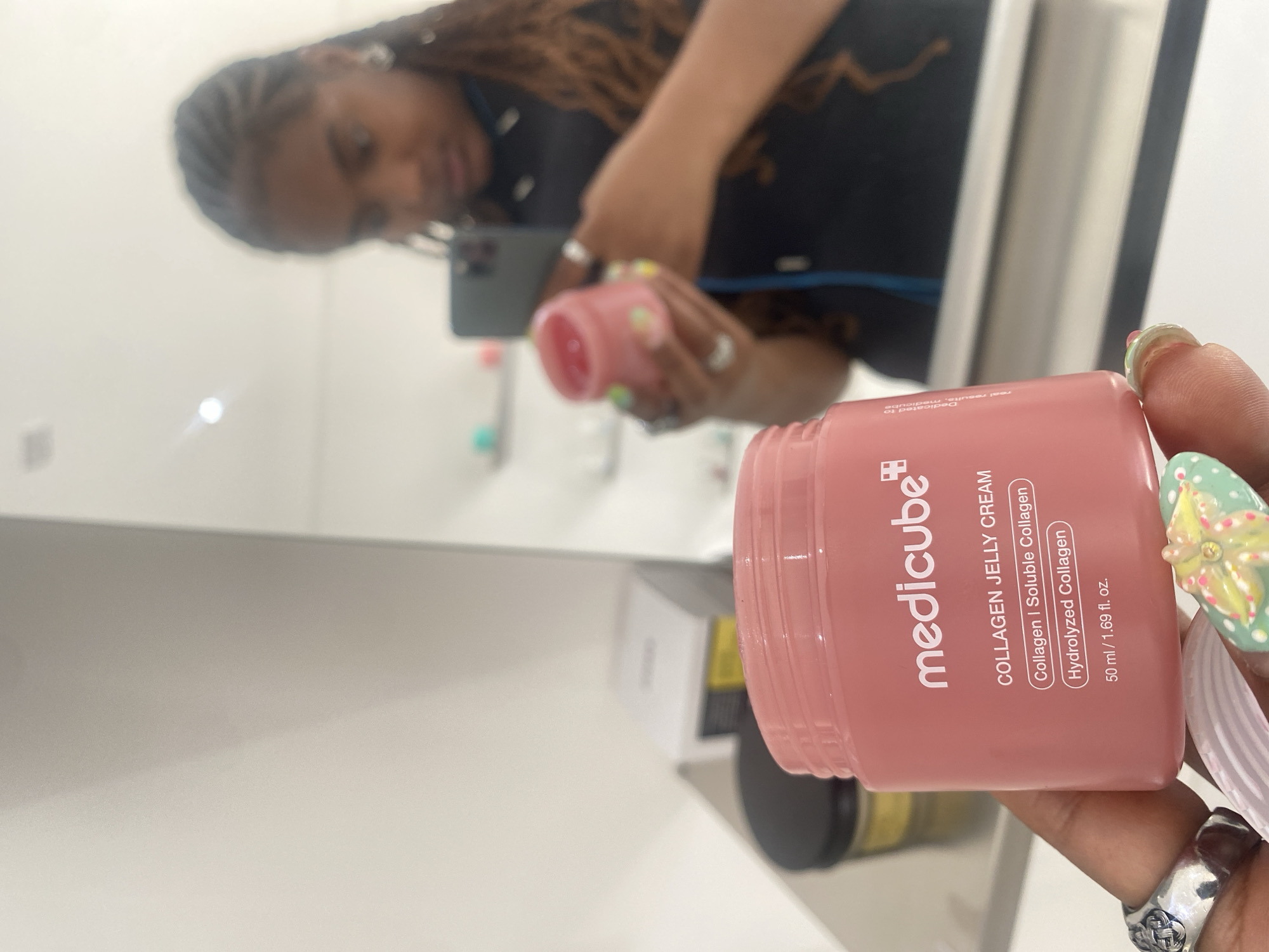 A person is holding a container of medicube Collagen Jelly Cream in front of a mirror. The container is pink and displays the product name and ingredients. The person is wearing multiple rings on their fingers and has decorative nail art. The mirror reflects the person holding a phone, possibly taking the picture. The background shows a white surface, potentially a countertop, with other cosmetic products visible. The image does not appear to be in London, United Kingdom.