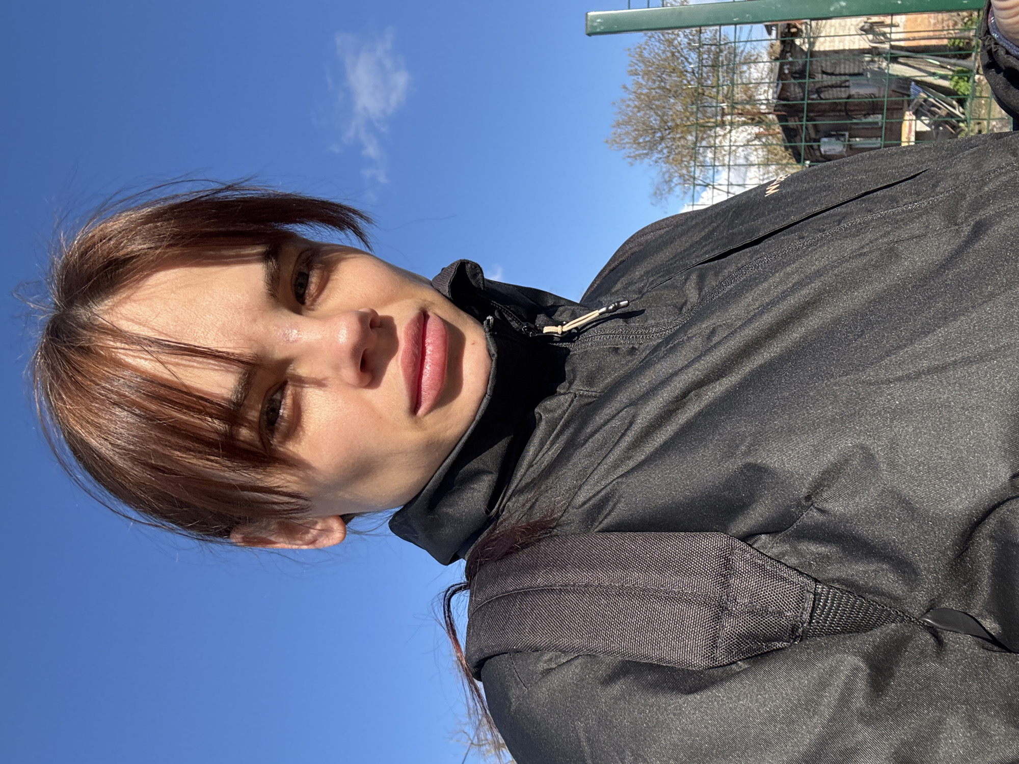 A photograph shows a person with dark hair and brown bangs wearing a black jacket and a black backpack. The background features a clear blue sky with a few clouds, a green metal fence, a tree, and a building. The image is oriented with the subject appearing to be lying down or looking upwards. No specific indicators of Rostov-on-Don are apparent.