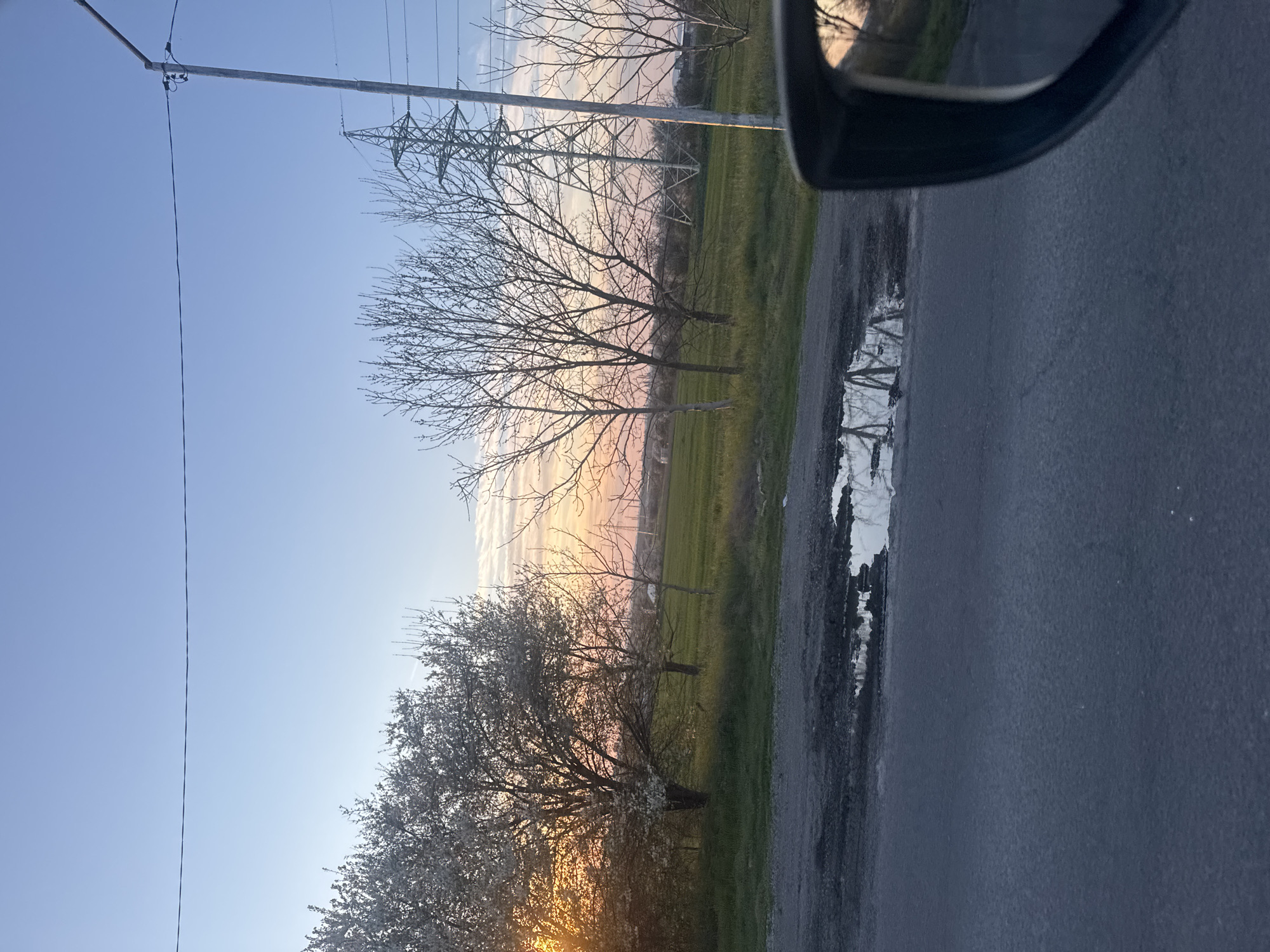 The image, likely taken from inside a vehicle, shows a roadside scene in Burgas, Bulgaria. A paved road occupies the foreground. To the right, a car's side mirror is visible. Puddles along the road's edge reflect the scene. Beyond the road is a grassy area interspersed with trees, some blossoming. Power lines and pylons rise in the background. The sky transitions from blue to a soft orange and pink, suggesting either sunrise or sunset.