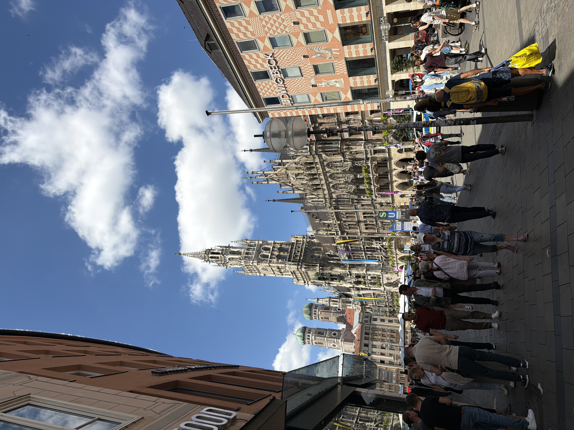 The image depicts a bustling street scene in Munich, Germany, likely in the city center. In the background, the iconic New Town Hall (Neues Rathaus) dominates the skyline with its intricate Gothic architecture and tall spire. Adjacent to it, the towers of the Frauenkirche (Church of Our Lady) are also visible, characterized by their green domes.
The street is paved with cobblestones and lined with buildings. On the left, a building with a reddish-brown facade and large windows is visible, with the word "FOOD" partially displayed on its side. To the right, a building with a distinctive patterned facade in shades of orange and white, featuring windows and the signage "LUDWIG BECK," stands prominently.
A crowd of people is walking along the street, suggesting a lively atmosphere. They are dressed in casual summer clothing, and their interactions are not clearly defined but indicate general pedestrian movement. Some individuals are carrying shopping bags, and a person in the foreground is wearing a yellow backpack. A police car is visible further down the street, and a bicycle is parked near the right edge of the frame. The sky is blue with scattered white clouds, indicating fair weather.