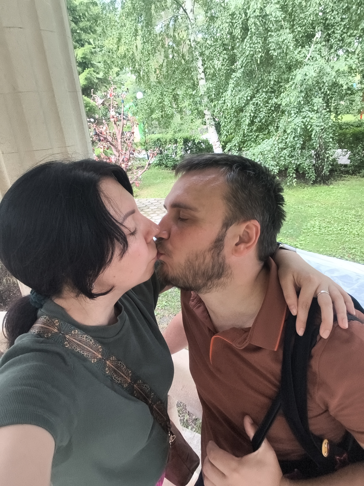A close-up, selfie-style, medium shot shows a man and a woman kissing outdoors, with lush green trees and foliage in the background. The woman, on the left, has dark hair pulled back with a green tie and wears a dark green t-shirt with an ornate, decorative band across the chest. Her eyes are closed. The man, on the right, has short dark hair and a beard, and wears a rust-colored polo shirt. He has his eyes closed as well, and his left arm is around the woman's shoulders, with her hand resting on his arm. A silver ring is visible on her left hand. A backpack strap is visible over the man's right shoulder. The background includes a partial view of a light-colored stone column on the left and a garden with decorative elements. The scene appears to be in a park or garden setting.