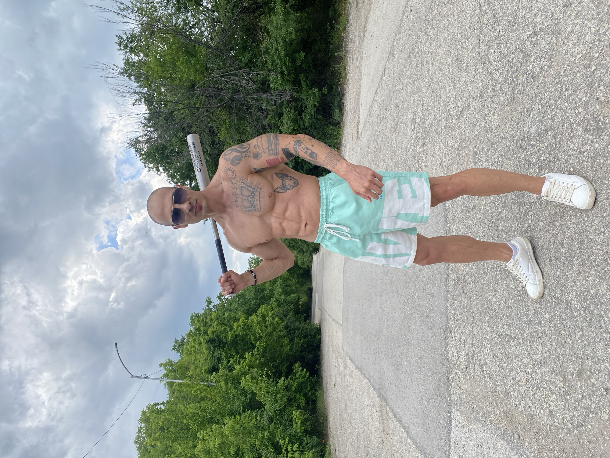 A shirtless, muscular man with tattoos stands on a paved road in an area with trees and an overcast sky, potentially in the vicinity of Pleven, Bulgaria. He wears sunglasses, white and light blue shorts, and white sneakers. He holds a baseball bat over his shoulder. In the background is dense green foliage and a utility pole. The ground is light-colored asphalt.