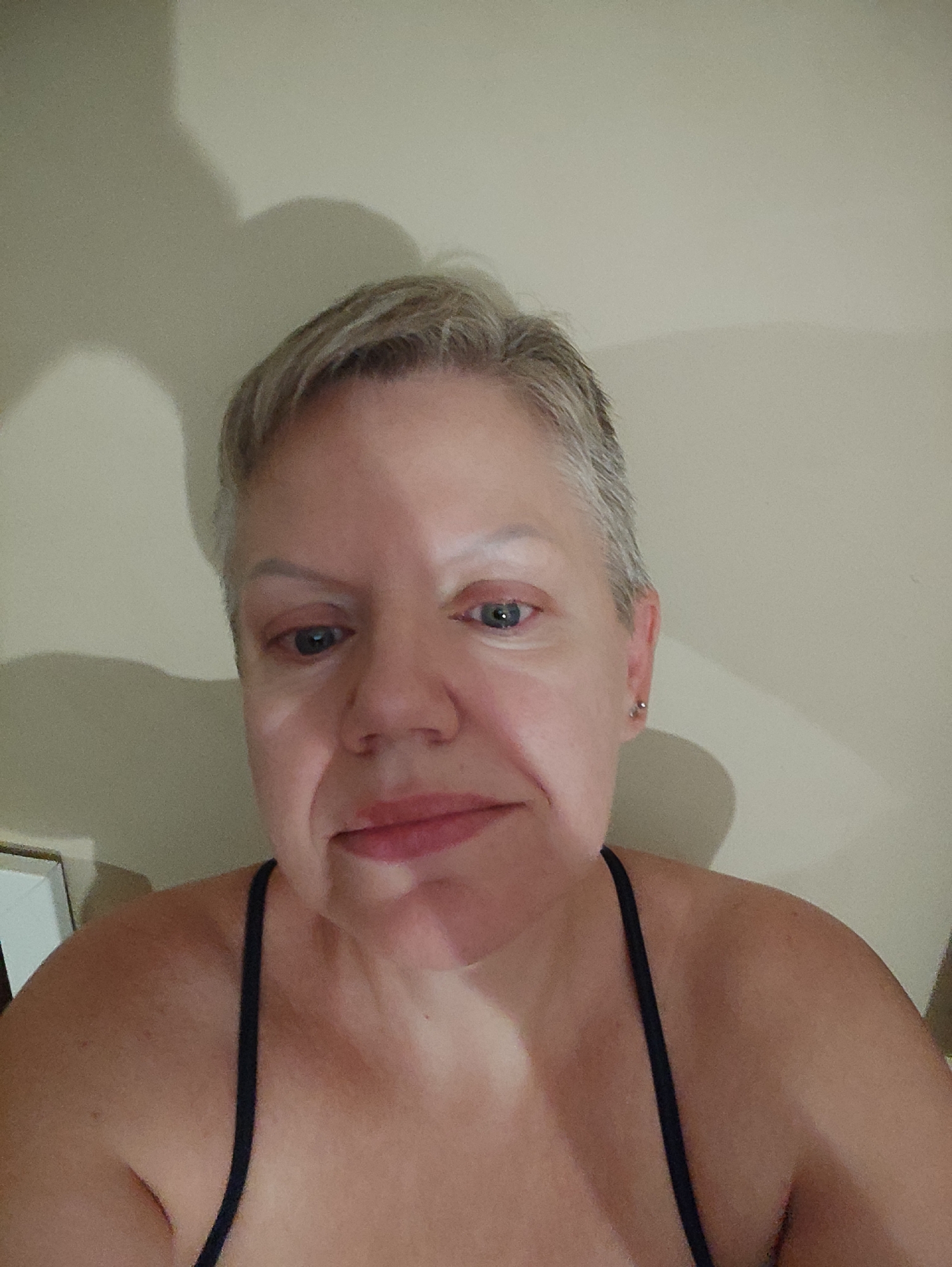 A close-up, head-and-shoulders selfie shows a woman with short, graying blonde hair and fair skin. She is looking directly at the camera with a neutral expression. Her eyes are open, and she is wearing a dark-colored tank top with thin straps. The background appears to be a plain, light-colored wall with a shadow cast on it from the left side of the frame. There is a hint of a shadow from the woman's head on the wall as well. The lighting is somewhat dim, creating a soft, diffused effect. The image does not provide any information about its specific location within Kadıköy, Turkey.