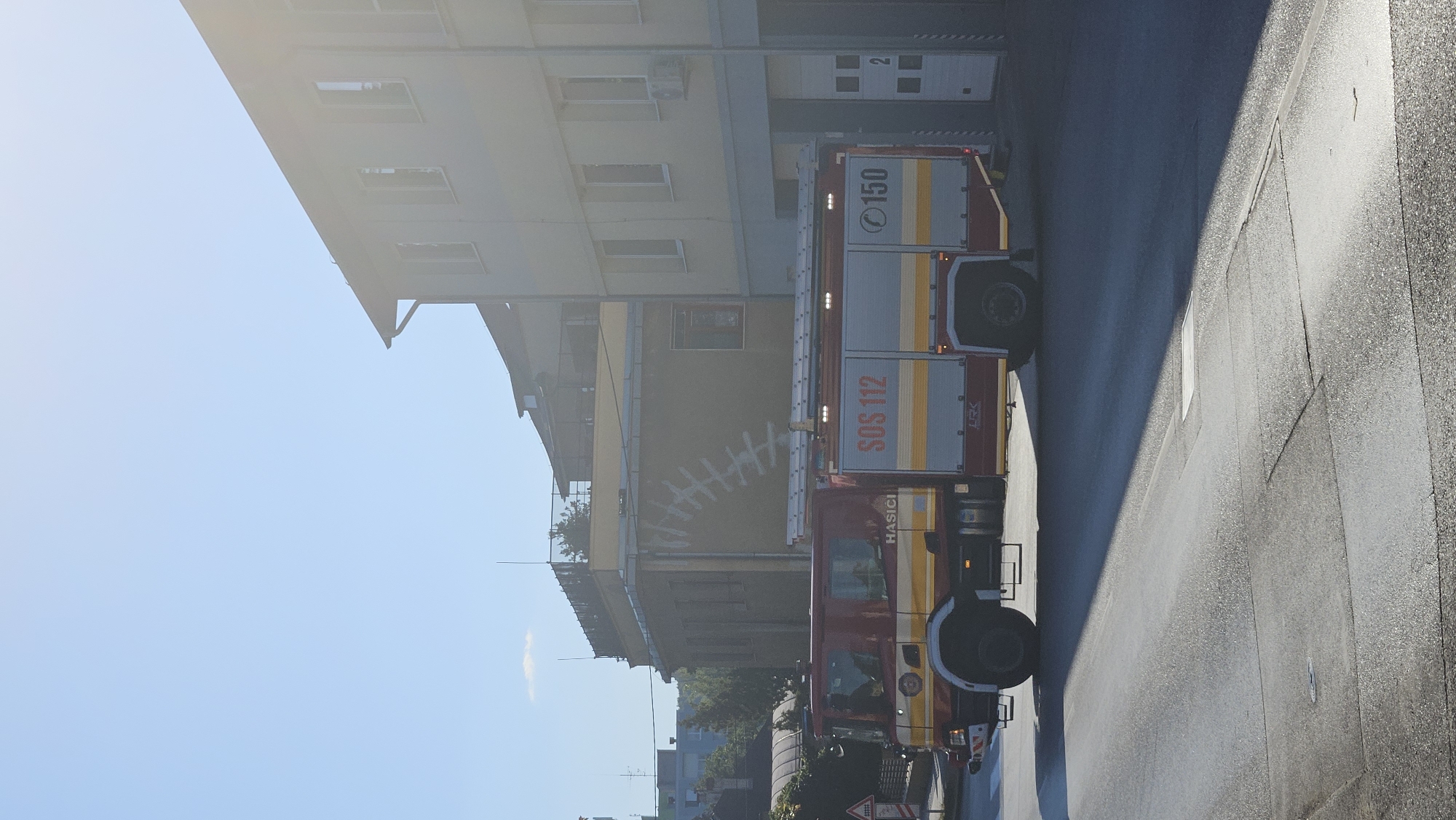 A side view of a red and yellow fire truck is captured in Prešov, Slovakia, parked on a paved road in front of a light-colored building. The fire truck has the word "HASIČI" printed on its side, which is Slovak for "firefighter." Above this, "SOS 112" is visible, indicating the emergency number. The number "150" is also present on the truck, likely representing another emergency contact or unit designation. The building behind the truck appears to be a fire station, with garage doors visible to the right. The sky is clear and blue, with the sun creating lens flare in the upper right portion of the image, casting a long shadow from the fire truck across the road. The overall impression is a typical urban scene with emergency services present.