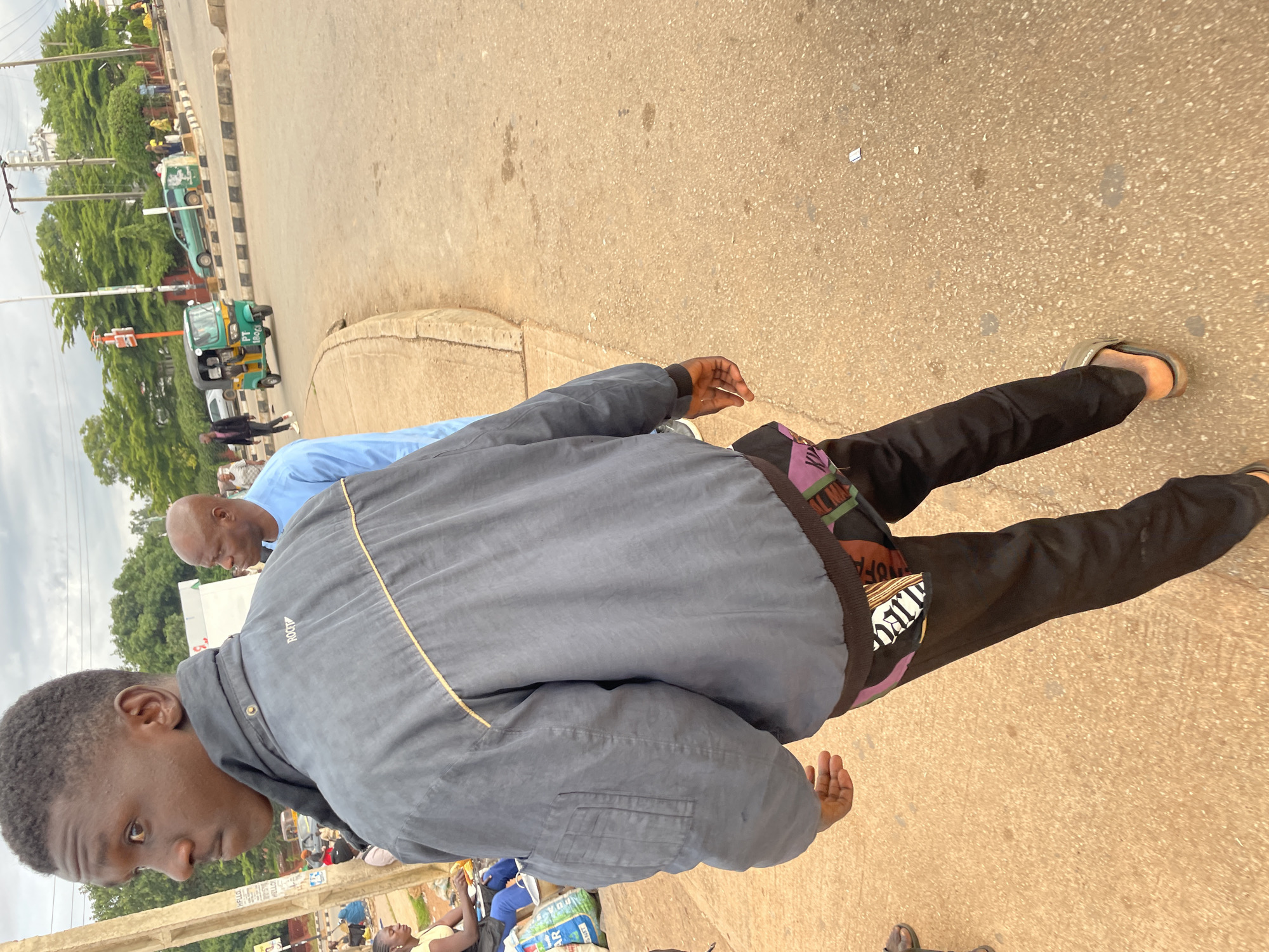 The image captures a street scene in Jos, Nigeria, likely during the daytime given the overcast sky. In the foreground, a young Black man wearing a dark gray jacket over black pants is walking away from the camera. The jacket has a gold trim detail across the back and a zippered pocket on the left sleeve. His pants have a colorful, patterned trim peeking out from the waistband. He is wearing open-toed sandals.
To his left and slightly behind him, another Black man with a shaved head is visible, wearing a light blue short-sleeved shirt. Further back and to the left, there are several people gathered, some appearing to be vendors or shoppers. A woman in a yellow top and blue pants is seated at a table with items on it, and a large bag of produce is on the ground next to her.
The background shows a busy street with vehicles, including a green and yellow tricycle or "keke" moving on the road. Other cars and a light green sedan are also present. A traffic light and streetlights are visible, along with trees and buildings in the distance. The ground is paved with reddish-brown asphalt and concrete. The overall impression is of a typical urban environment in Jos.