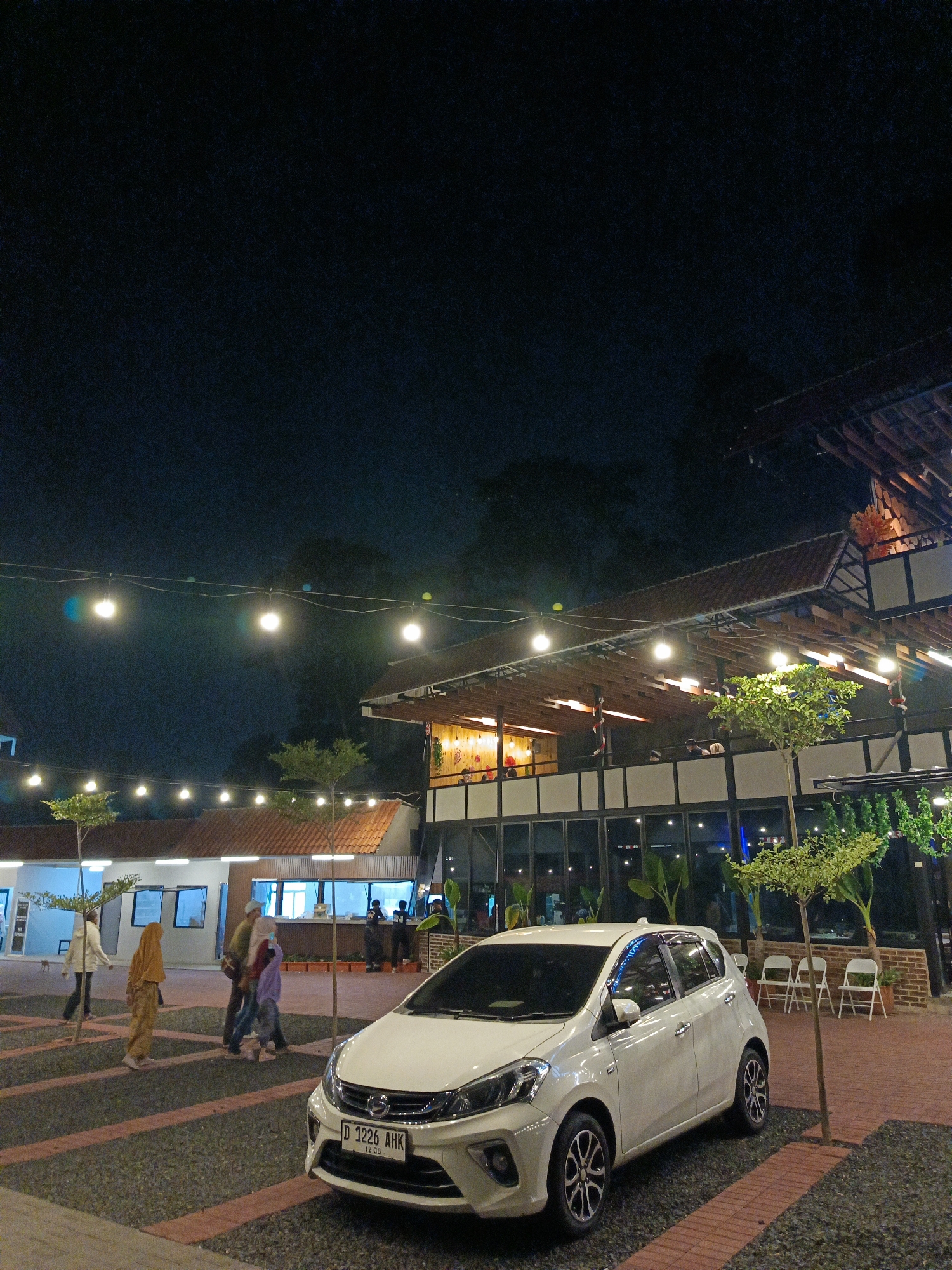 The image depicts an outdoor area at night, likely a commercial establishment in Cimahi, Indonesia, with a white car parked in the foreground. The car, a Perodua Axia or similar model, has its license plate visible, displaying "D 1226 AHK". Behind the car, there is a paved area with gravel and brick accents. Several people are walking in the midground, including two women dressed in hijabs and two children. One woman is wearing a beige hijab and matching outfit, and another is wearing a purple hijab. They appear to be walking together.
In the background, a modern building with large glass windows and a distinctive roof structure is illuminated by string lights. The architecture suggests a restaurant or cafe. The building has multiple levels, and some patrons can be seen inside and on an elevated balcony. Several small trees are planted around the area, and white folding chairs are visible near the building. The sky is dark, indicating nighttime. The overall ambiance is that of a lively commercial district at night.