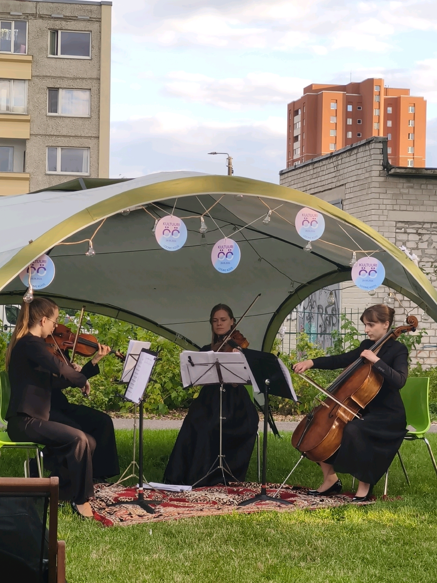 Three musicians are performing under a green canopy in a grassy outdoor area. Two women are playing violins, and one woman is playing a cello. They are all dressed in black. There are music stands in front of them. In the background, there is a brick building and a tall orange building. The event appears to be part of "Kultuuri Loomu Koos" as indicated by the circular decorations hanging from the canopy. The weather is overcast with visible clouds.