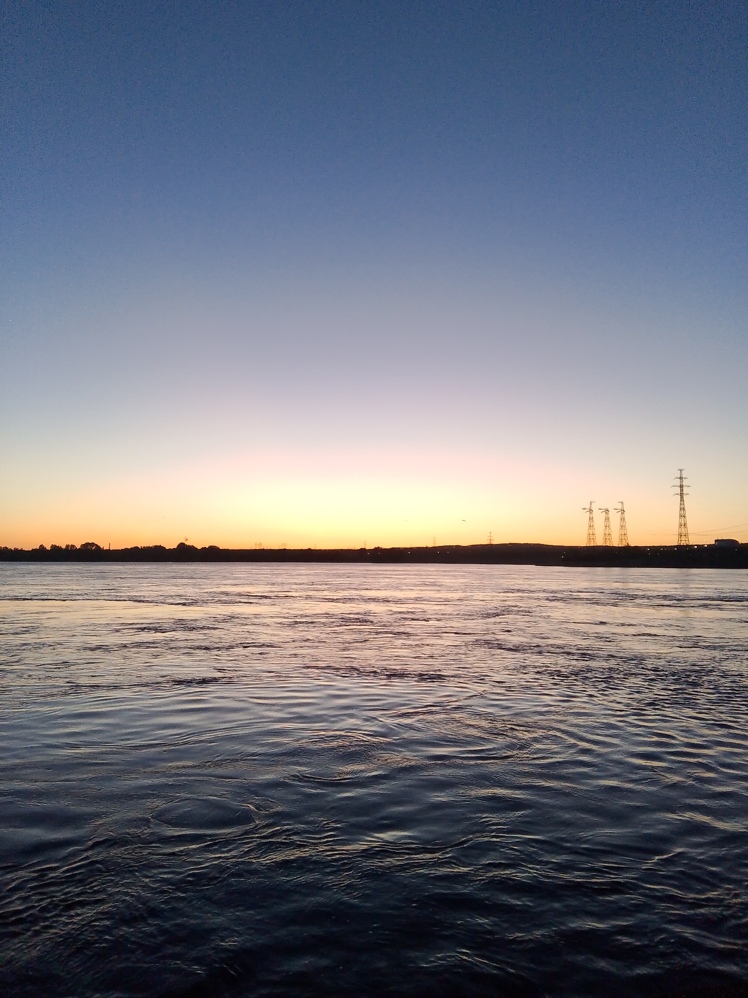 The image depicts a wide body of water, possibly the Kama River, reflecting the colors of a twilight or sunset sky in Chaikovsky, Russia. The sky transitions from a deep blue at the top to a light orange/yellow horizon. Silhouetted against the horizon are trees and what appear to be electrical transmission towers. The water surface is disturbed by ripples. No people are visible.