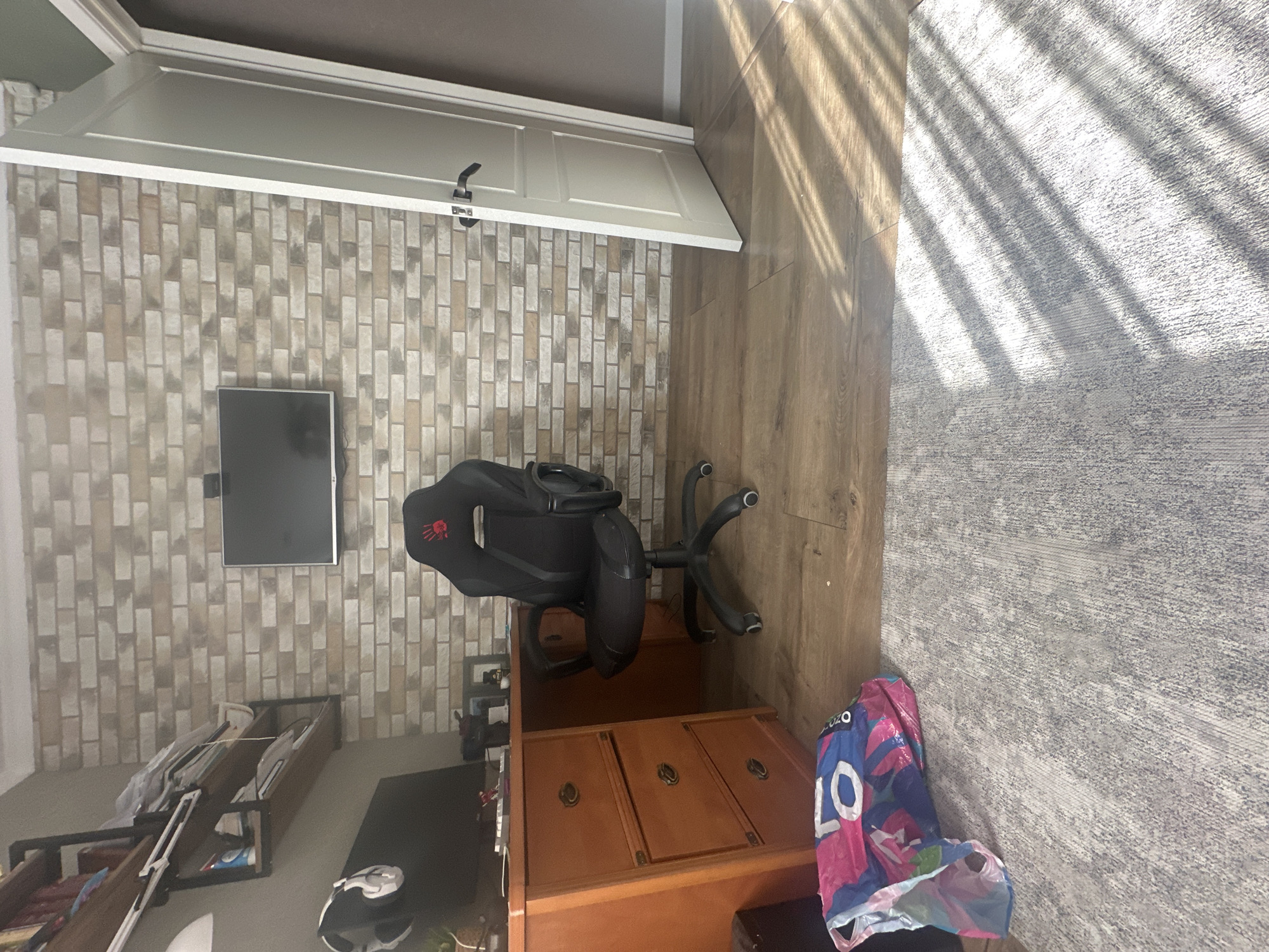 The image shows a room in Unknown, Unknown. The room has a light-colored brick wall with a mounted TV and shelves holding various items. A black gaming chair is positioned in front of a wooden desk with drawers. A shopping bag rests on the floor next to the desk, on a light-colored rug. The floor is made of wood-patterned planks. Sunlight is streaming in through a window, casting shadows on the floor and rug. A white door is visible at the top of the wall.