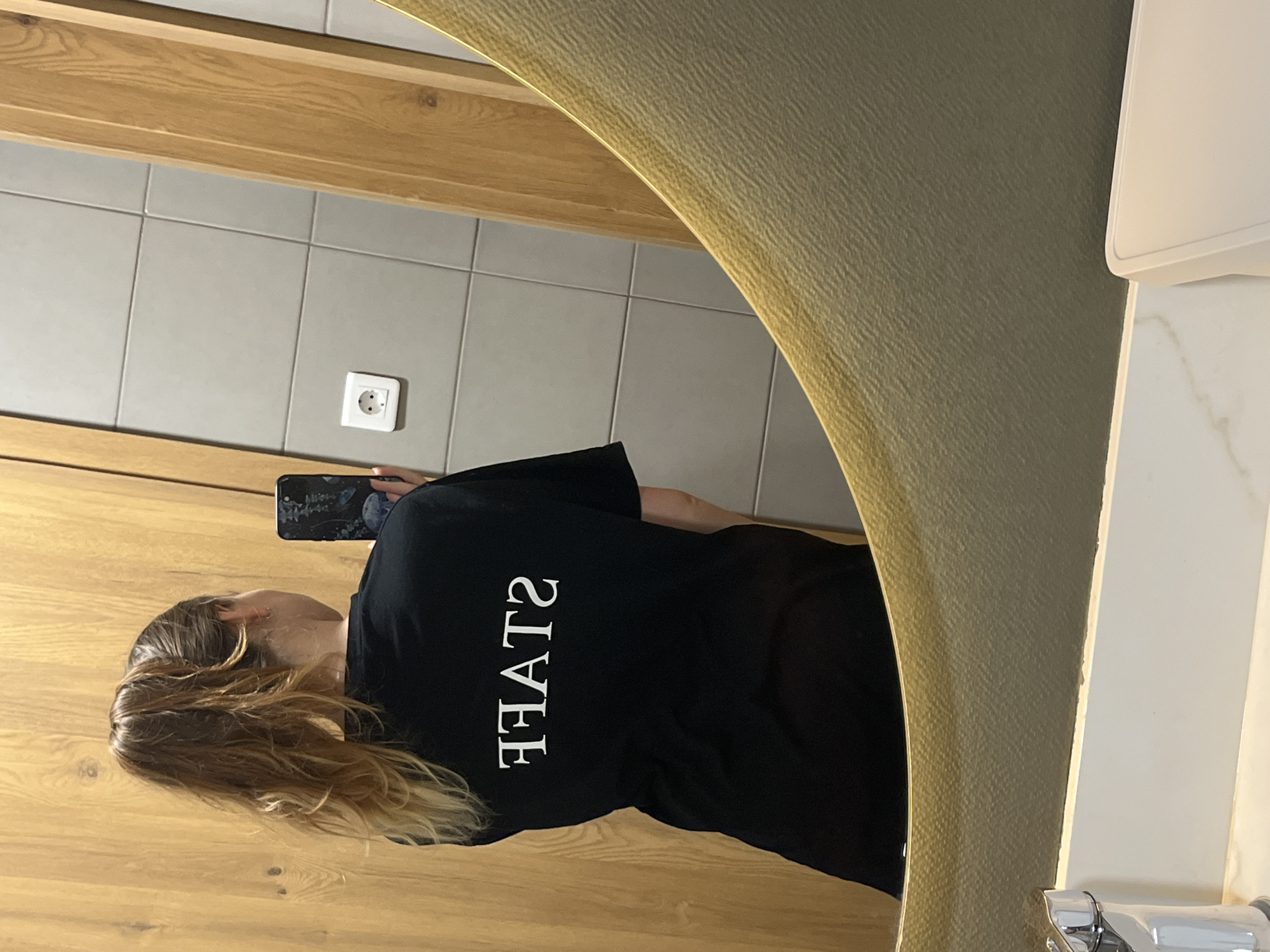 The image shows a woman from the back, wearing a black t-shirt with white text that reads "STAFF" in reverse. She has her hair pulled up in a high ponytail. She is holding a smartphone in her right hand, which is facing away from her, suggesting she is taking a selfie in a mirror. The background consists of a tiled wall, a wooden door, and a sink with a chrome faucet. The scene appears to be in a bathroom or a similar personal space, possibly in Plovdiv, Bulgaria, given the context of the request. The lighting is moderate, and the overall image quality is good.