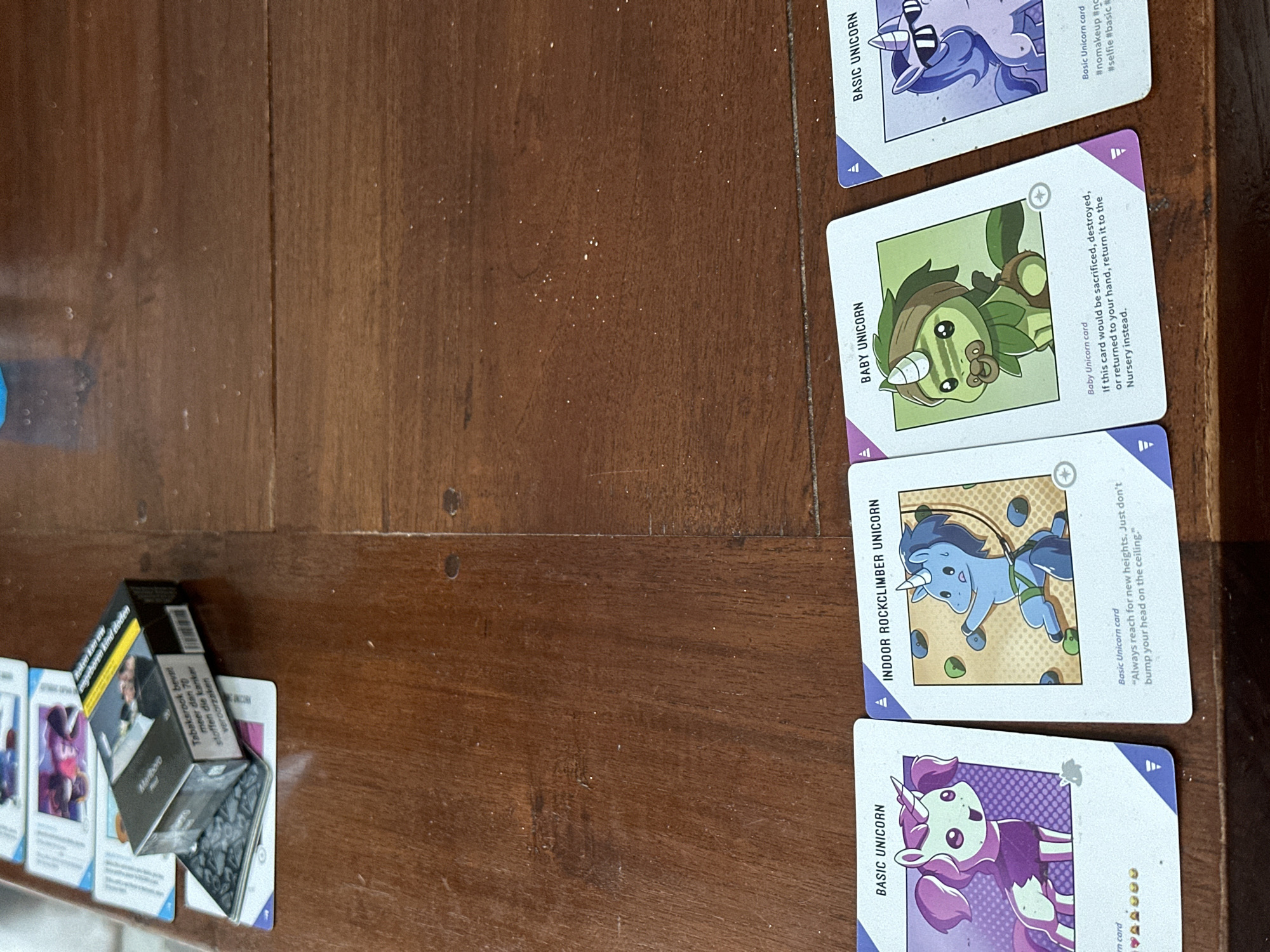 The image shows a wooden table with playing cards and a pack of cigarettes. The playing cards appear to be from the card game "Unstable Unicorns" and feature cartoon unicorns. The pack of cigarettes is a Marlboro brand and has Dutch text printed on it. The location is not discernible from the image, and there is no evidence that it is in Amsterdam, Netherlands.