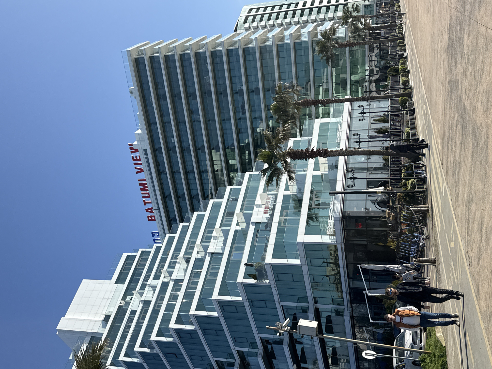A daytime, high-angle shot captures a modern urban scene in Batumi, Georgia, featuring a distinctive high-rise building with a clear blue sky as its backdrop. The prominent building, emblazoned with the red text "BATUMI VIEW" vertically along its side, showcases a unique stepped or zig-zag architectural design with extensive glass facades that reflect the sky and surrounding elements. Behind this building, another modern high-rise is visible, also featuring numerous glass windows.
A wide, paved promenade or street runs parallel to the buildings, lined with tall palm trees that cast light shadows. Several individuals are present on this paved area. In the foreground, two men are clearly visible, one wearing a dark jacket and jeans, and the other in a light-colored vest over a white shirt and jeans. Further down the promenade, other people and what appear to be bicycles or scooters are discernible. Street lamps with multiple ornate lights are spaced along the pavement. A white car is partially visible near the bottom left, obscured by a pole.
The scene is brightly lit by the sun, indicating clear weather. Reflections on the glass buildings show glimpses of other structures and the vibrant blue sky. A small, less distinct sign on one of the building's windows appears to say "FOR SALE OR RENT." The overall impression is one of a contemporary coastal city environment during a sunny day.