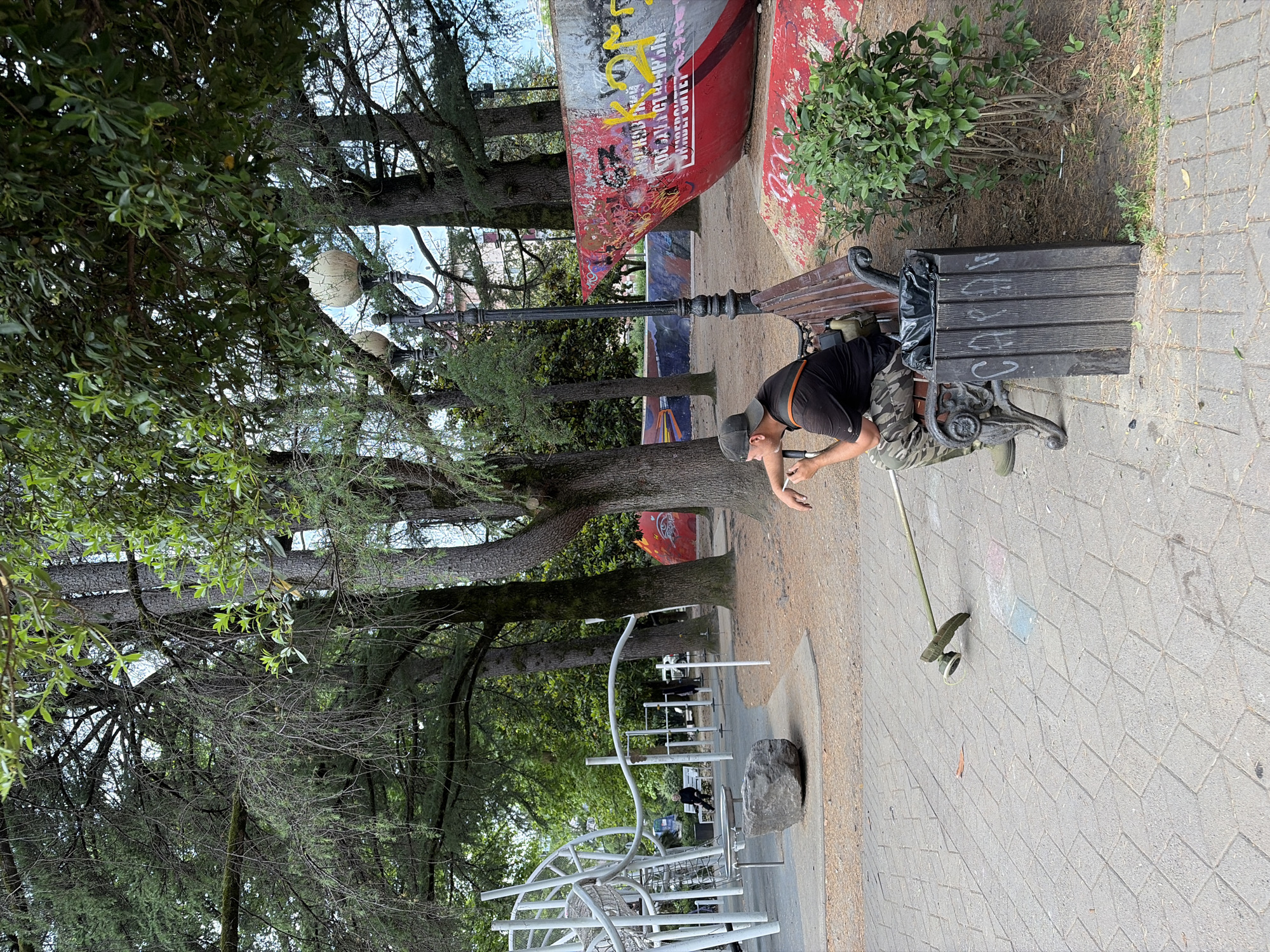 The image depicts a man sitting on a park bench in Sochi, Russia, with a string trimmer next to him. The man is wearing a black t-shirt, camouflage pants, and a baseball cap. He is holding a cigarette and appears to be taking a break from yard work. The park is set with trees, a playground, and a large red and silver structure covered in graffiti. The ground is paved with grey tiles and there is a trash can next to the bench. The overall atmosphere suggests a casual, outdoor setting within a public space.