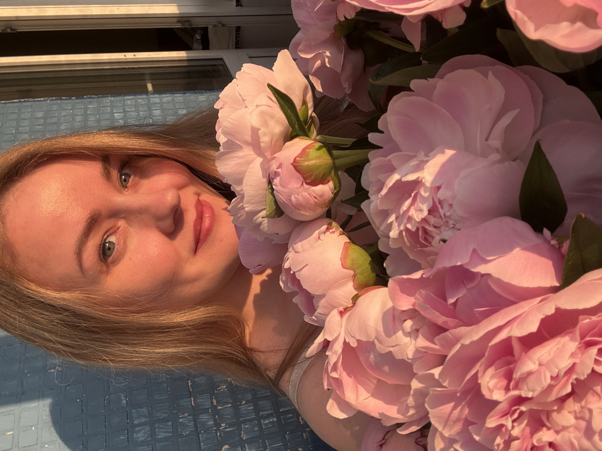 A close-up, head-and-shoulders portrait of a young Caucasian woman with fair skin, blonde hair, and blue eyes, holding a large bouquet of pink peonies. She is wearing a light-colored, possibly white, spaghetti-strap top. The lighting suggests it is late afternoon or early evening, with warm sunlight illuminating her face and the flowers. The background consists of a textured blue wall with a distinct pattern of rectangular tiles, and a sliver of a white door frame or window frame on the right. A shadow of the woman's head and shoulders is cast on the blue wall to her left. The overall impression is one of gentle, natural beauty and warmth, possibly captured outdoors on a balcony or patio in Chaikovsky, Russia, given the blue tiled wall reminiscent of some older Russian apartment buildings and the soft, golden hour light.