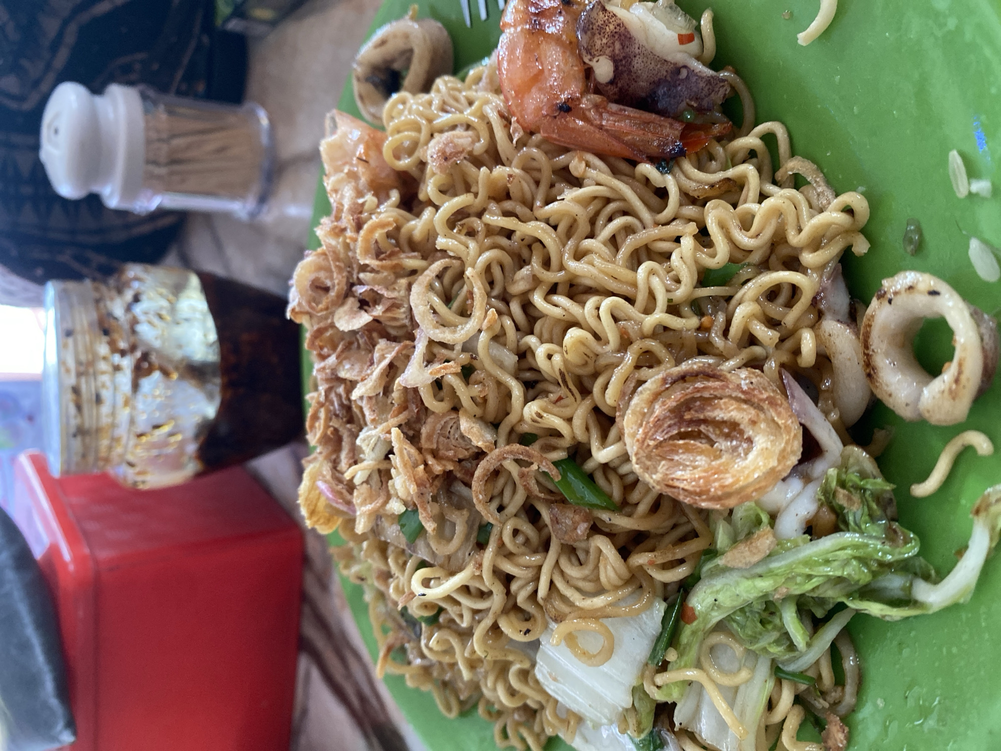 This close-up image showcases a vibrant plate of stir-fried noodles, likely Mi Xao, served on a bright green, round plate. The golden-brown, wavy noodles are generously topped with an array of ingredients. Prominently featured are several large, reddish-orange grilled or pan-fried shrimp, some still with their shells, and tender white calamari or squid rings.
A significant portion of the noodles is covered with an abundant scattering of crispy, golden-brown fried shallots or onions, providing a textural contrast. Mixed within the noodles are pieces of blanched or stir-fried green vegetables, possibly Chinese cabbage, along with chopped green onions, adding freshness and color. The dish appears well-seasoned and appetizing, suggesting a flavorful meal.
To the left of the plate, various table items are visible. A transparent shaker filled with wooden toothpicks and topped with a white plastic lid stands upright. Below it, a glass jar containing a dark, reddish-brown oily condiment, possibly chili oil or a spicy sauce, rests on a bright red, rectangular base or container. The tines of a metallic fork are partially visible, protruding from the noodles at the top left. The background is softly blurred, revealing hints of a patterned surface and a lighter table, indicating a casual dining setting, consistent with an eatery in Tien Thanh Ward, Vietnam. The scene captures the moment a meal is presented, ready for consumption. There is no visible text in the image.