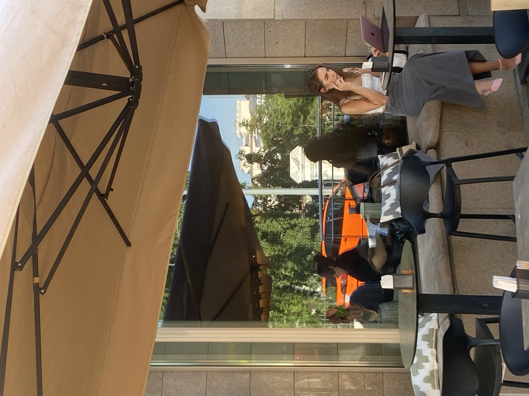 The image shows an outdoor cafe scene, likely in Kazan, Russia, as the reflection in the window shows a building that resembles Kazan architecture. A woman is sitting at a table with a laptop. She appears to be talking on the phone. The table is partially covered by a large umbrella. Through the window, one can see a street scene with trees and a reflection of an orange vehicle. Several tables and chairs are visible, suggesting an outdoor dining area. The cafe appears to be located next to a sidewalk.
