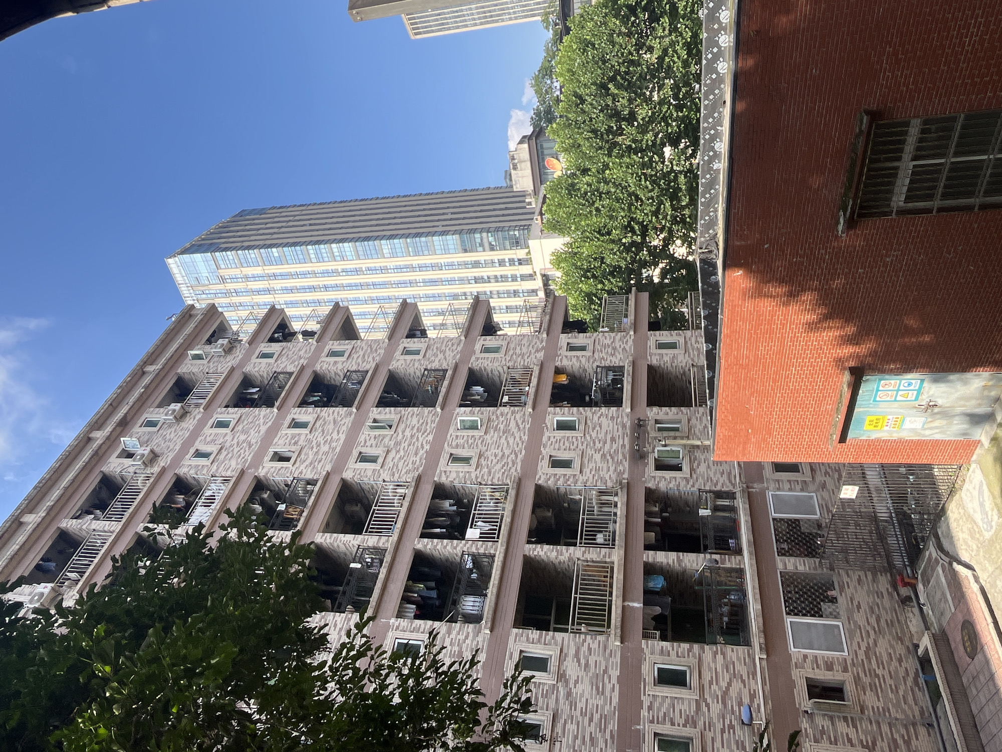 This image shows a dense urban scene in Yuzhong District, China, on a clear, sunny day. In the foreground, a red brick building occupies the lower right portion of the frame, with a large window visible. To the left of this building, an apartment complex with a patterned brick facade is prominent. This complex has multiple floors, each featuring balconies with railings. Laundry is visibly hanging from clotheslines on many of these balconies, suggesting residential activity. Several air conditioning units are mounted on the exterior walls of the apartment building. In the background, two modern skyscrapers rise above the apartment complex. One is a tall, glass-fronted building, and another is a more rectangular skyscraper. Lush green trees are interspersed between the buildings, adding a touch of nature to the urban landscape. The sky above is a clear, bright blue with a few wispy clouds. The overall impression is one of a bustling city with a mix of older and modern architecture.