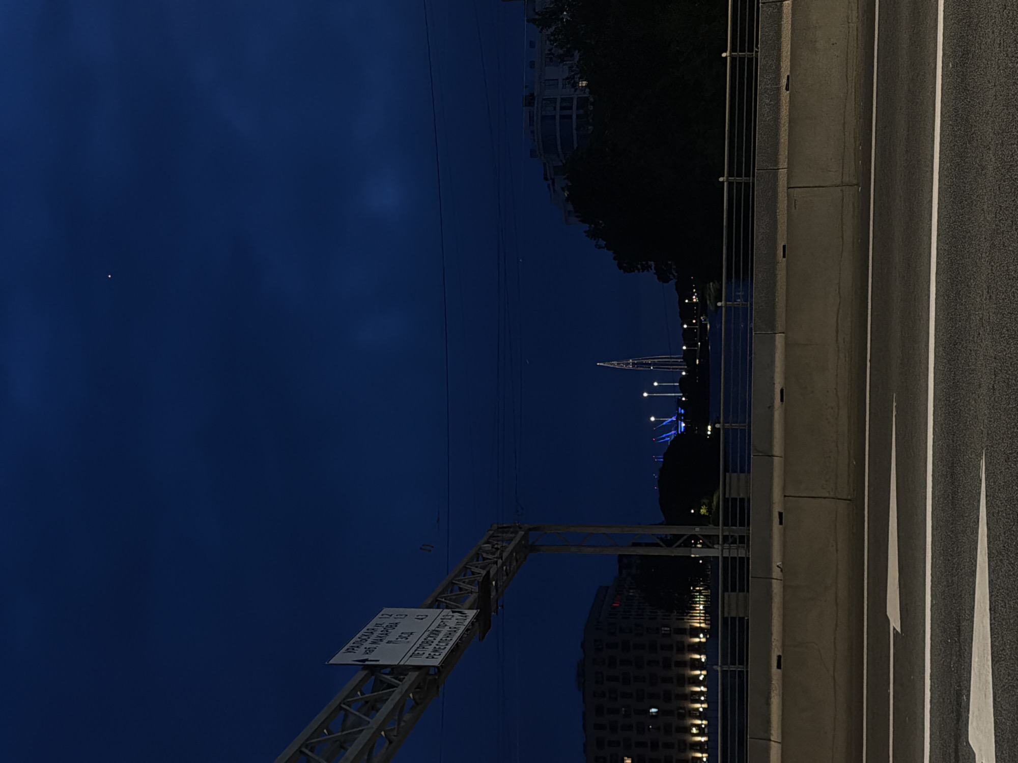 The image is a nighttime photograph taken from a road. In the foreground, there is a concrete barrier and asphalt road with white directional arrows. In the middle ground, there is a body of water with a railing along its edge. Buildings with illuminated windows are visible on the left bank of the water. On the right bank, there is a dark silhouette of trees and a tall, slender structure that appears to be a skyscraper or tower. In the distance, illuminated structures and lights are visible. The sky is dark blue, with a small bright star or planet visible in the upper right quadrant. A road sign with text in Cyrillic is attached to a metal gantry on the left side of the image. The overall scene depicts an urban environment at night.