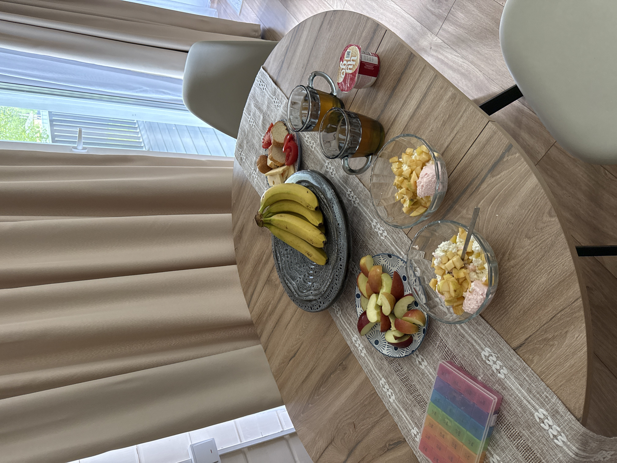 The image shows a breakfast scene indoors, likely in a residential setting. A round wooden table is the focal point, laden with breakfast items arranged on a beige table runner with a repeating pattern. The food items include two glasses of orange juice, a container of yogurt, slices of bread and tomato, a bunch of bananas on a grey plate, sliced apples on a blue and white patterned plate, and two bowls of what appears to be fruit salad with a dollop of pink cream. A spoon is visible in one of the fruit salad bowls. A multi-colored pill organizer sits near the edge of the table. Two chairs, one with a tan-colored back and one white, are positioned around the table. A window with light beige blinds is visible in the background. The location information (Buzhaninovo, Russia) cannot be confirmed from the image alone.