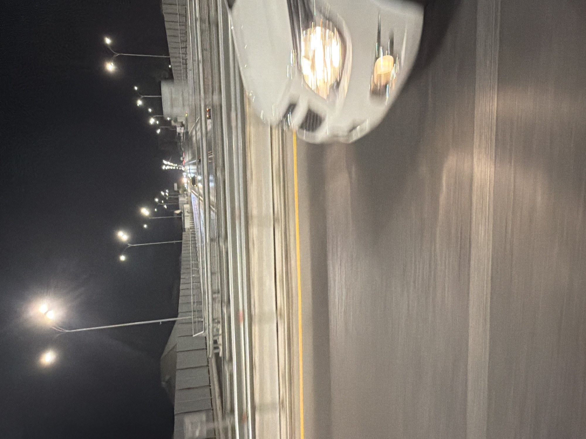 The image depicts a night scene on a highway, likely in or near Tashkent, Uzbekistan, characterized by blurred motion due to the camera's movement. The foreground is dominated by the right side of a white car, its headlights illuminated and appearing blurred. The road surface is dark asphalt with visible horizontal motion streaks. A bright yellow line marks the lane divider.
In the mid-ground, a series of streetlights on tall poles line the highway, casting a glow in the dark sky. A concrete sound barrier, segmented by vertical supports, runs along the left side of the highway. Beyond the barrier, a large, indistinct structure, possibly a building or an overpass, is visible.
The background shows multiple lanes of traffic, with the lights of other vehicles appearing as streaks of red and white. The highway appears to be an elevated structure or a bridge, with railings and structural elements visible along its edges. The overall impression is one of a busy highway at night, with the movement of vehicles and the artificial lighting creating a dynamic visual effect.