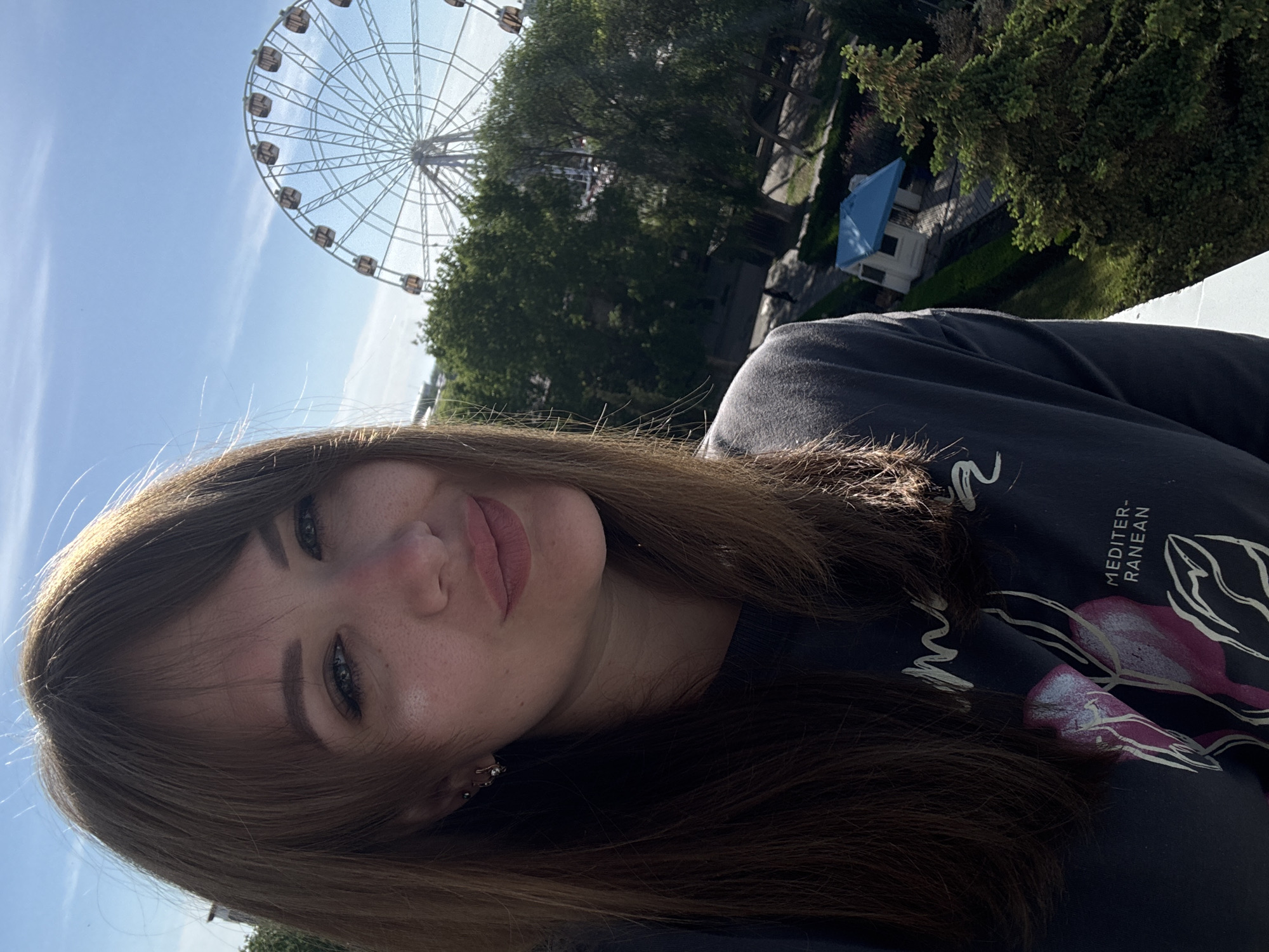 A selfie captures a young woman with brown hair, fair skin, and dark eyes looking directly at the camera with a slight smile. Her hair is parted to the side, and she wears a dark-colored shirt with a graphic design.
In the background, a large Ferris wheel is visible against a partly cloudy, blue sky. Green trees surround the Ferris wheel, suggesting a park or recreational area. A small, light-colored structure with a blue roof is also visible amid the trees.
Based on the presence of the Ferris wheel and similar architectural styles, the location is likely the Yevpatoriya Park of Culture and Recreation in Yevpatoriya, Ukraine.