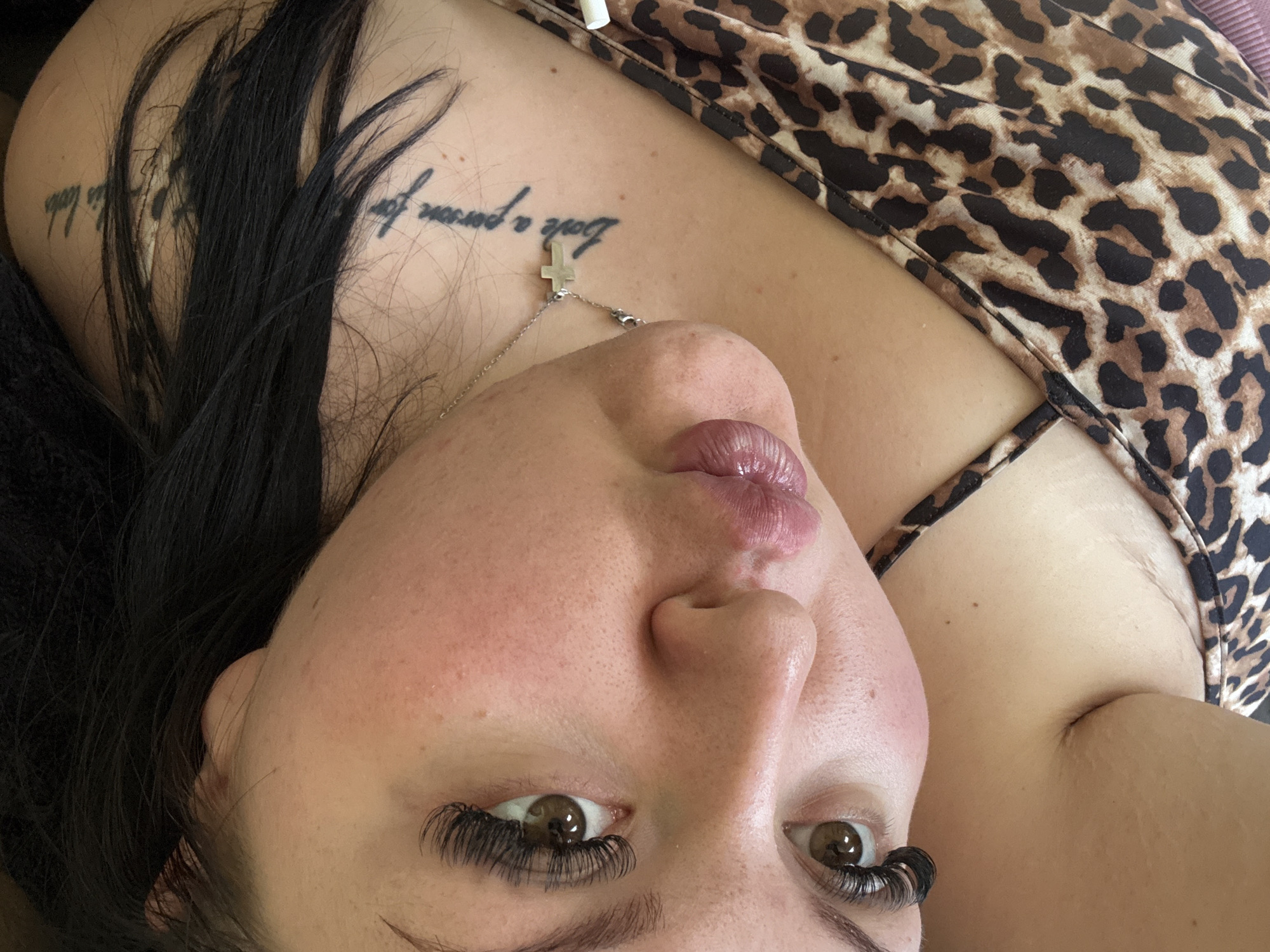 A close-up, angled view of a person, likely female, with fair skin. The person has dark hair and long, dramatic eyelashes. Their eyes are brown. They are wearing a necklace with a small cross pendant. A tattoo is visible on their chest that reads "Love a person for..." which may be incomplete or partially obscured. They are wearing an animal print top. The location in Sofia, Bulgaria is not discernible from this image.