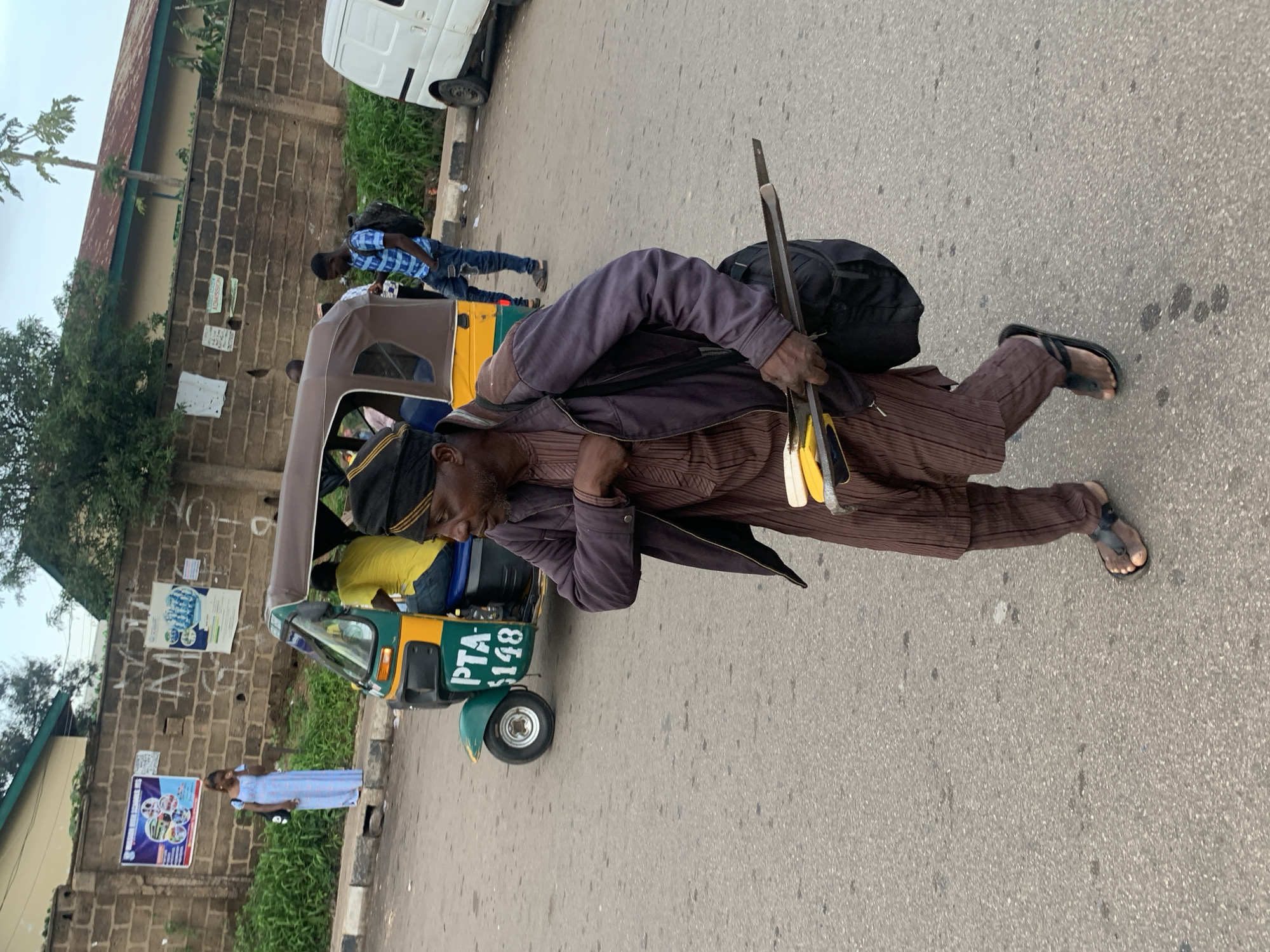 In Barakin Lerawi, Nigeria, a man walks across a paved road, carrying a long saw and a bundle of yellow objects, possibly wooden dowels, slung over his left shoulder in a black bag. He wears a brown, pinstriped tunic and trousers, a dark skullcap, and black sandals. In the background, a green and yellow auto-rickshaw, labeled "PTA $148," is parked, with passengers visible inside. A white van is parked to the right. A brick wall, adorned with posters, forms the backdrop, with lush green foliage bordering the sidewalk. The sky is overcast.