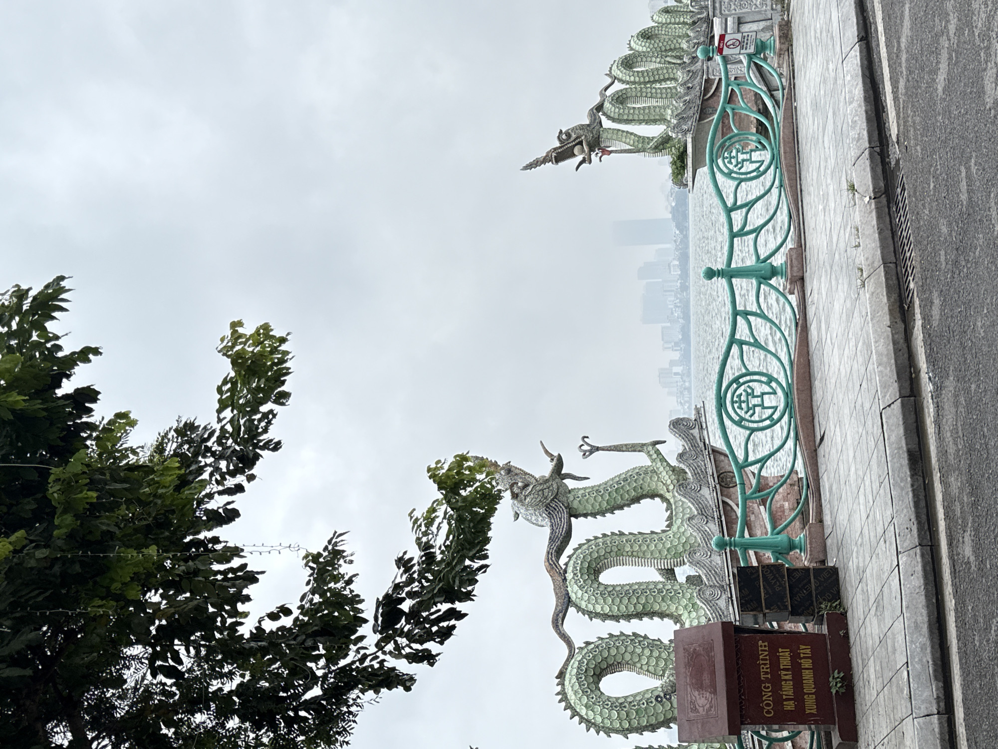 The photograph depicts a section of a pedestrian walkway in Hà Nội, Vietnam. A decorative green railing with coiled dragon sculptures lines the walkway, separating it from the water. A sign in Vietnamese is affixed to the railing. A tree with dense green foliage is visible to the left. In the background, across the water, is a cityscape shrouded in a hazy or overcast sky. The walkway is paved with light-colored stone tiles and is adjacent to a gray road surface. The overall light is muted, suggesting an overcast day.