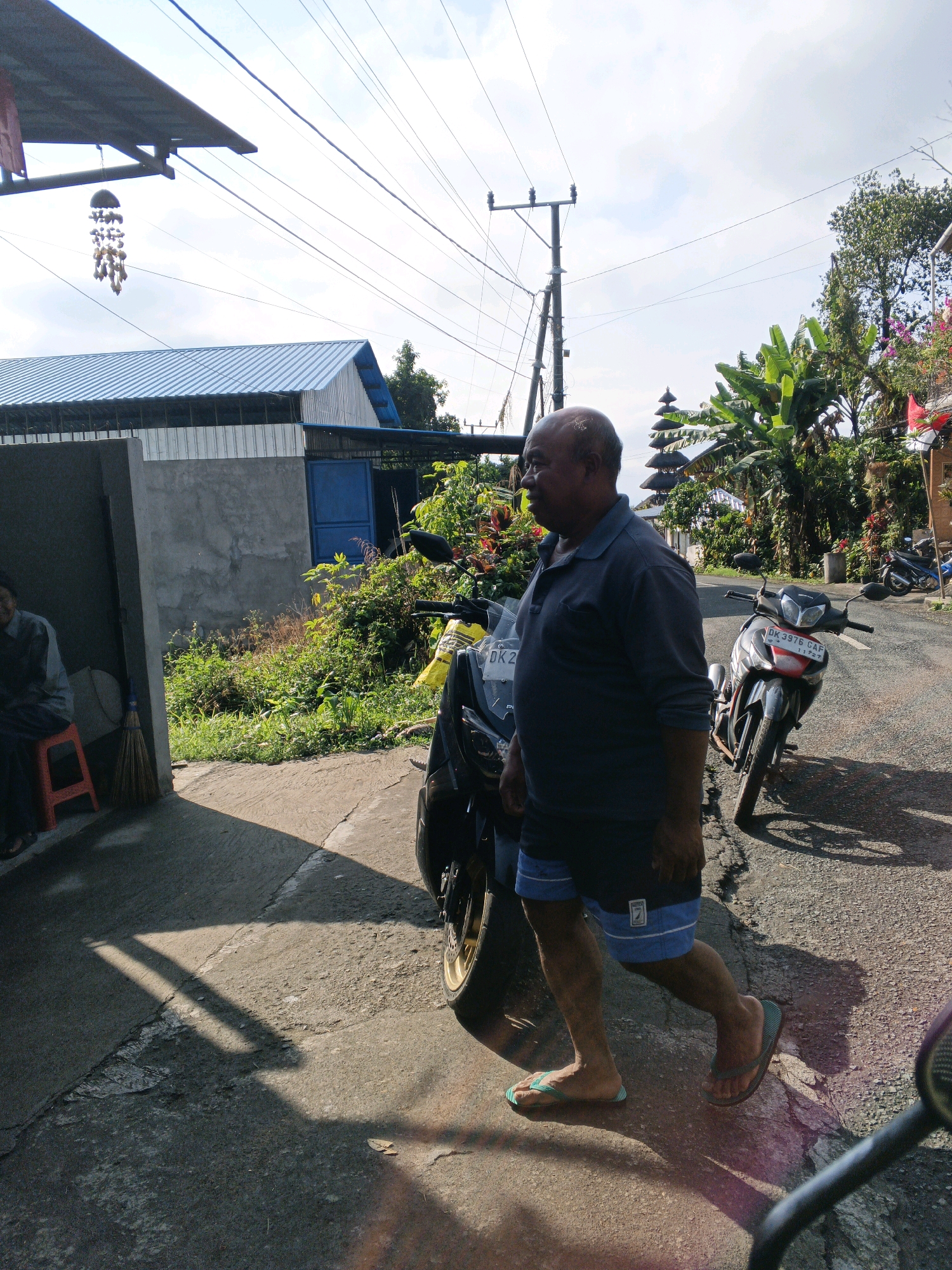 An outdoor, eye-level, medium shot shows an overweight, bald, middle-aged man with brown skin wearing a dark blue polo shirt and blue shorts walking from left to right in front of a motorcycle parked on the side of a paved road. The man's left leg is bent, and his right leg is extended as he walks. He is wearing green flip-flops on his left foot and brown flip-flops on his right foot. The motorcycle in the foreground is dark gray with a red and white license plate. The man is looking to his left. Behind him, further down the road, another motorcycle is parked. The scene is located in Penebel, Indonesia. The road is lined with lush green vegetation, including banana trees. In the background, a multi-tiered temple structure is visible. The sky is bright and partly cloudy. A structure with a blue roof and gray walls is on the left side of the image, with a person sitting on a red stool inside. A broom leans against the wall. The sun casts long shadows on the ground.